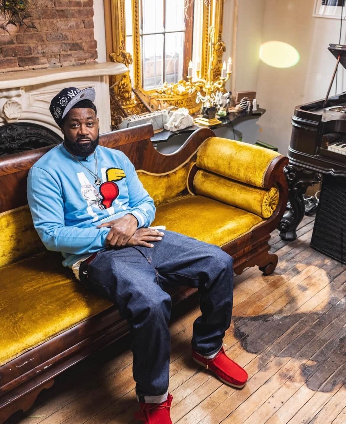 A man sitting on a vintage yellow velvet and wood sofa in a decorated room with a fireplace, ornate mirror, and a piano.