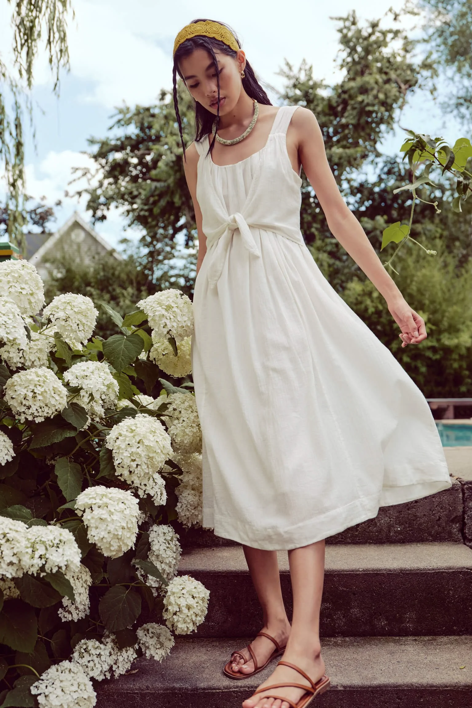 A young woman in a white sleeveless dress and brown sandals stands on stone steps beside a bush with white hydrangea flowers, outdoors with trees and a cloudy sky in the background.