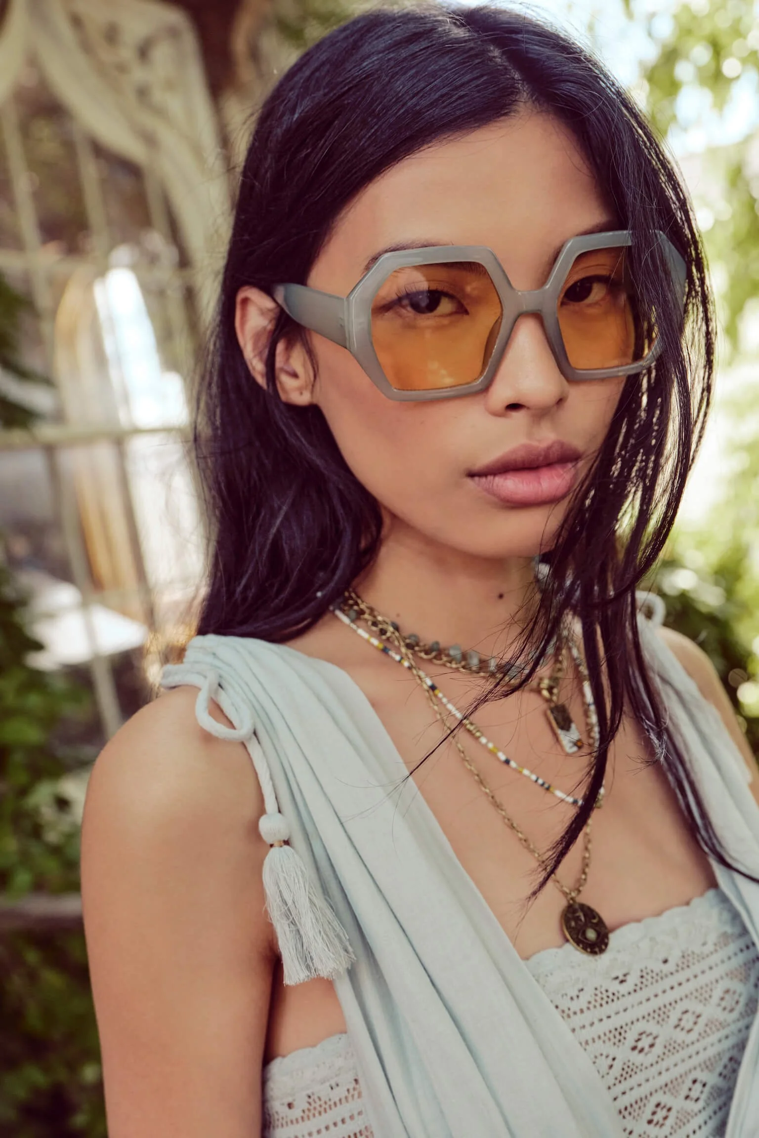 A young woman with black hair wearing large orange-tinted sunglasses and layered necklaces, posing outdoors with blurred greenery in the background.