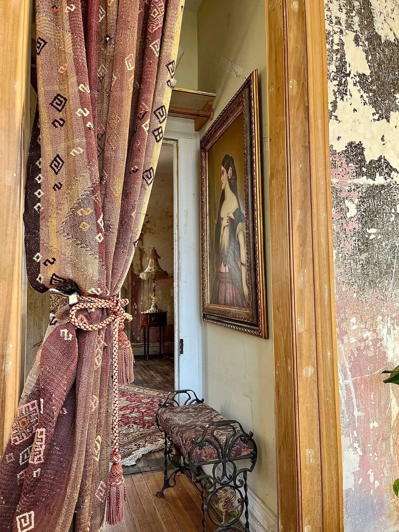 A vintage hallway with an ornate framed portrait of a woman hanging on a wooden-paneled wall. To the left, a patterned curtain is tied back, revealing a doorway to another room with a small table and a lamp. The hallway floor is wooden, and there is 