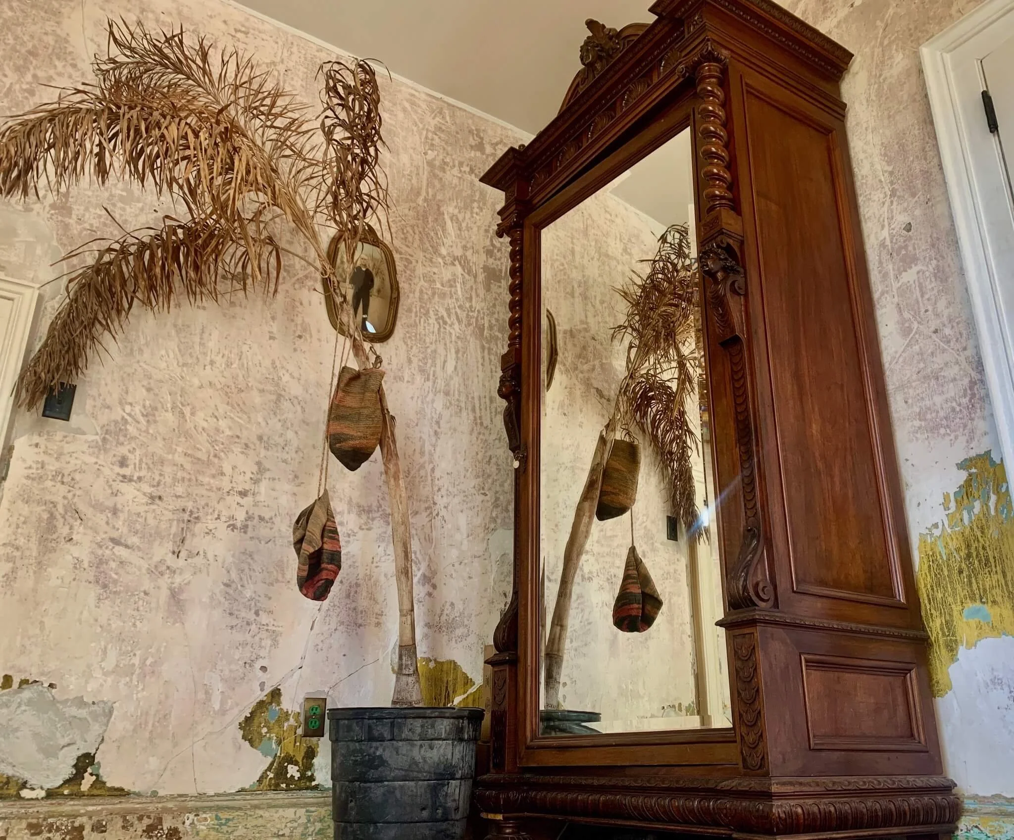 An antique wooden cabinet with a mirror reflecting a dried palm tree in a pot, hanging on a distressed wall with peeling paint. There is a wall picture and a black electrical outlet near the base of the wall.