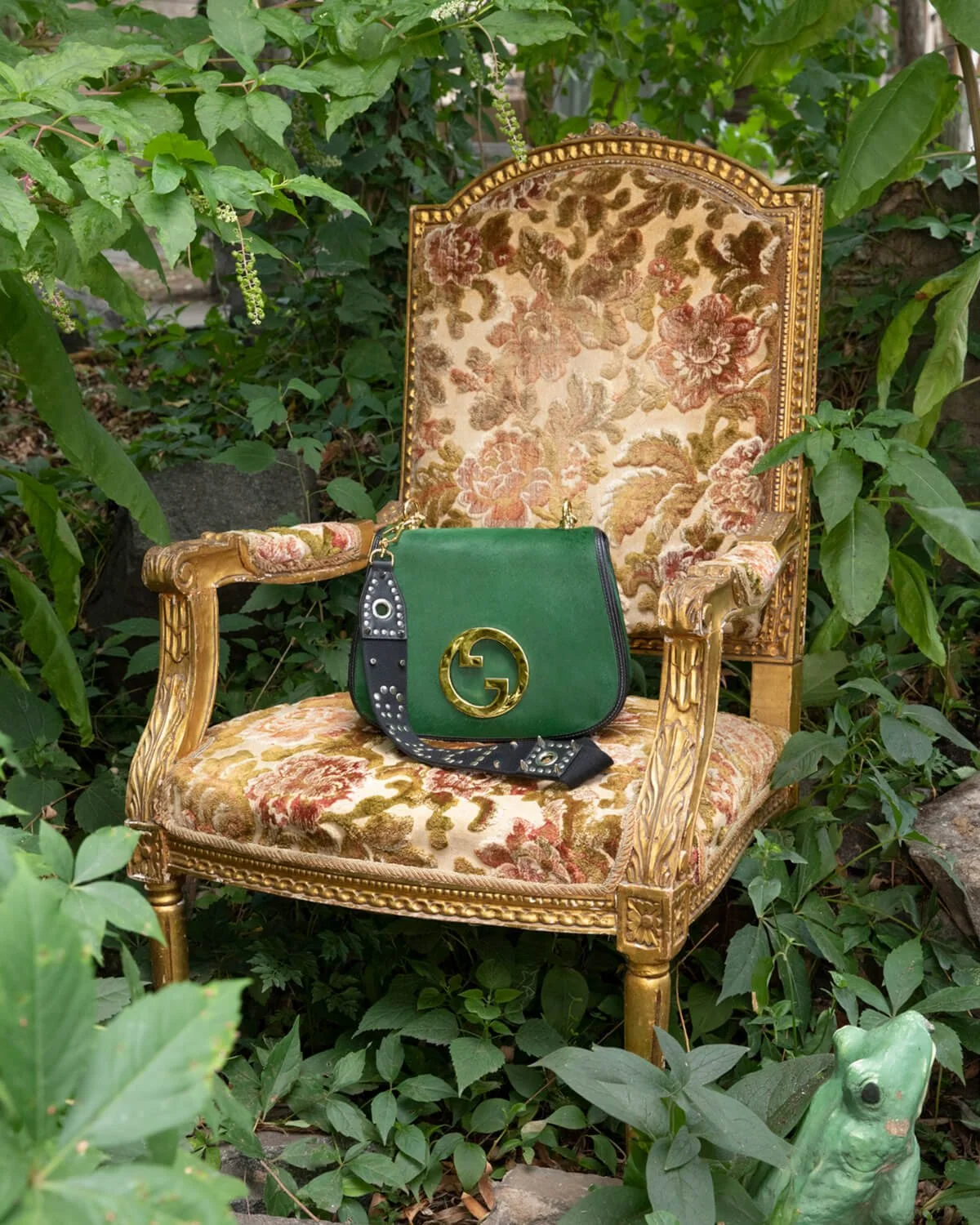 A vintage-style ornate gilded armchair with floral upholstery surrounded by green foliage, with a green Gucci handbag with a black strap placed on the seat.