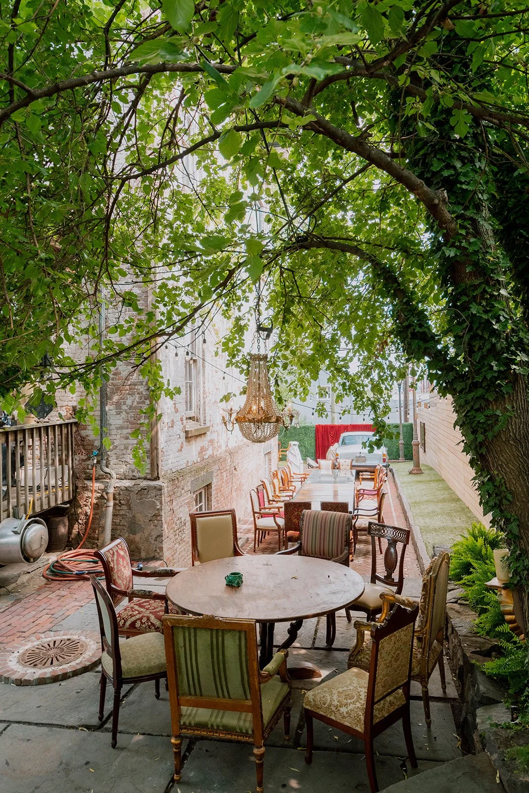 Outdoor patio area with vintage chairs surrounding a round table, hanging chandelier, and a vintage car in the background amidst greenery and brick walls.