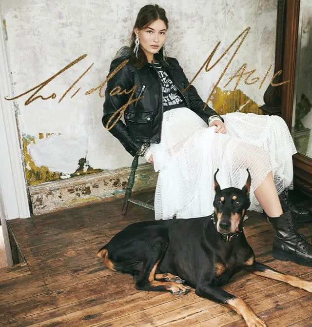 A woman with dark hair and hoop earrings sitting on a wooden floor with a black and tan dog lying beside her. She is wearing a black leather jacket, a black graphic T-shirt, a white skirt, and black boots. The background features a worn wall with pee