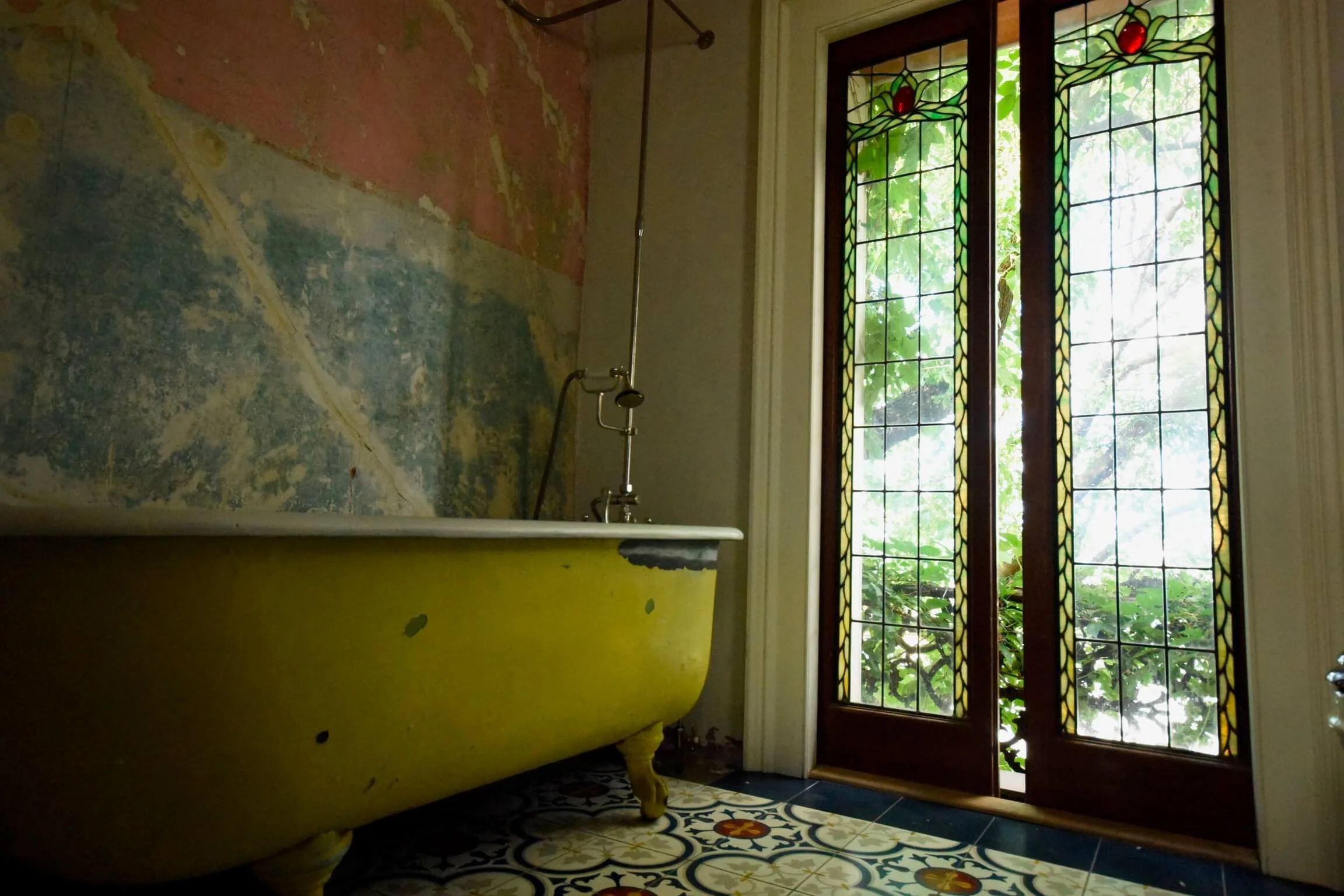 Vintage yellow clawfoot bathtub in a bathroom with textured wall paint, patterned tile floor, and stained glass door leading outside.