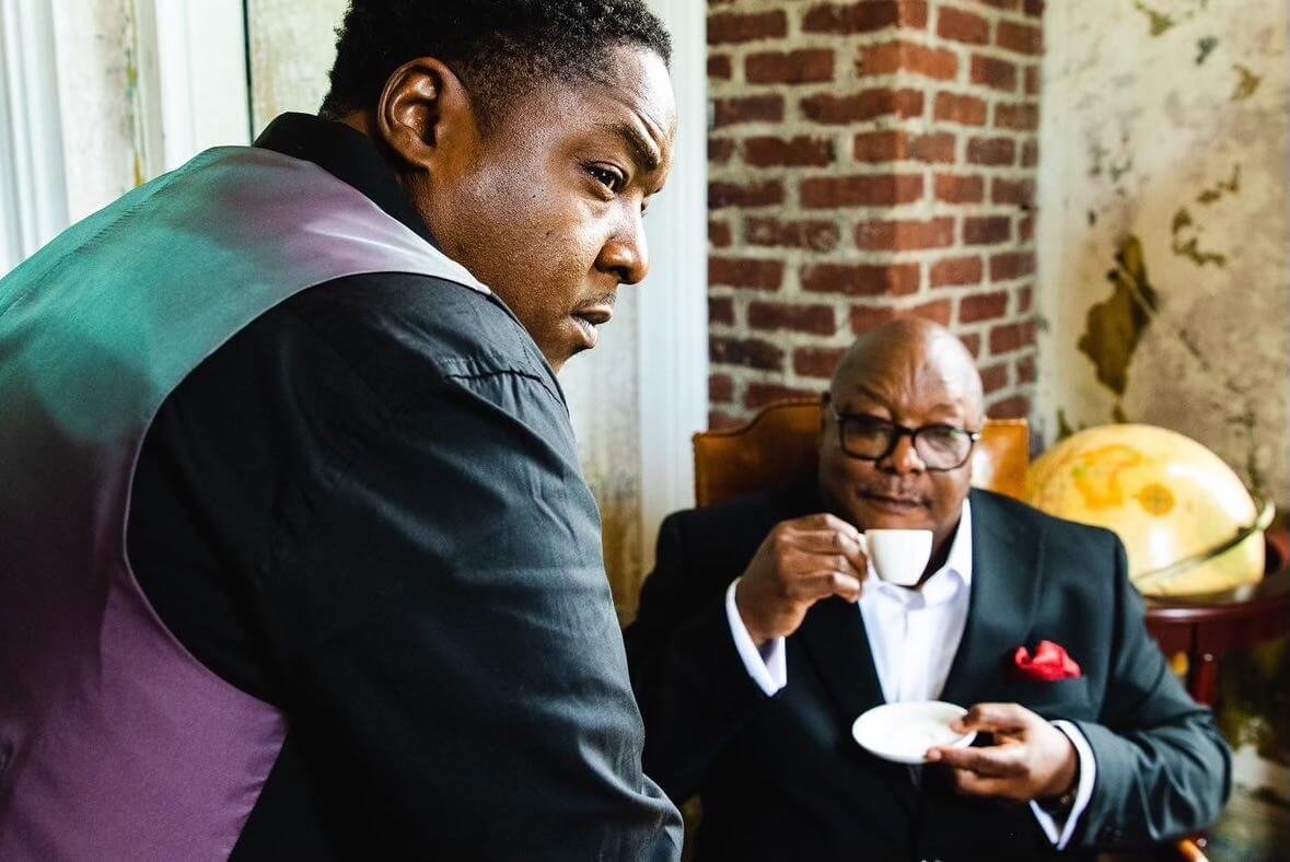 Two men having tea in a room with exposed brick wall and peeling plaster, one standing and one sitting in a suit with glasses, holding a teacup and saucer.