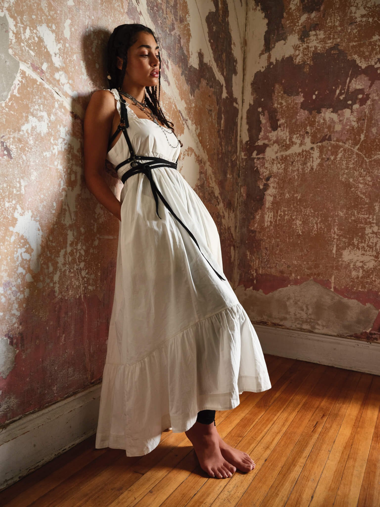 A woman with dark hair styled in braids wearing a long white dress with black straps and chains, standing barefoot against a textured, worn wall with peeling paint.