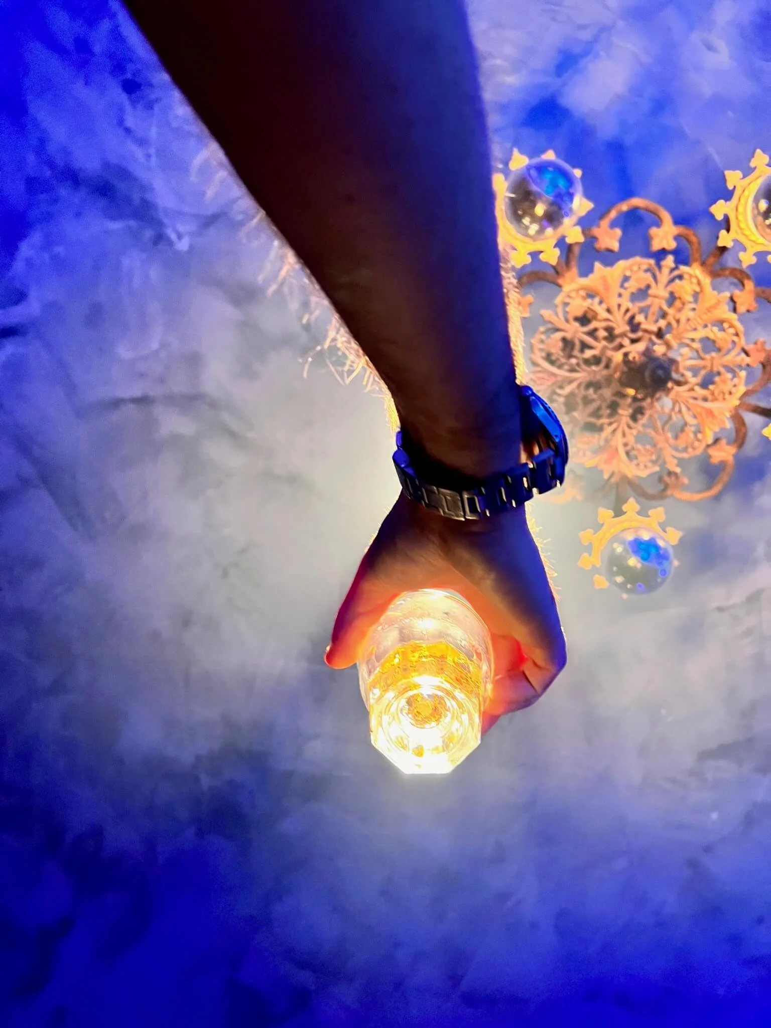 A person's hand holding a lit candle against a blue ceiling with ornate chandelier and decorative ornaments.