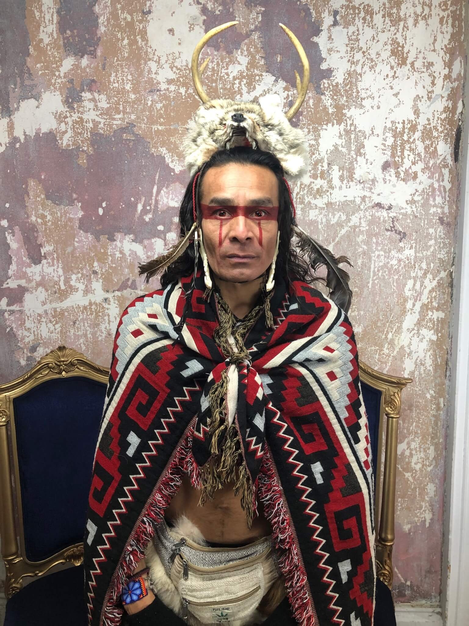 A man sitting on a gold and blue chair, dressed in traditional Native American-inspired attire, with face paint and a blanket. He wears a headdress made of a wolf skull with deer antlers and fur. The background is a weathered, painted wall.