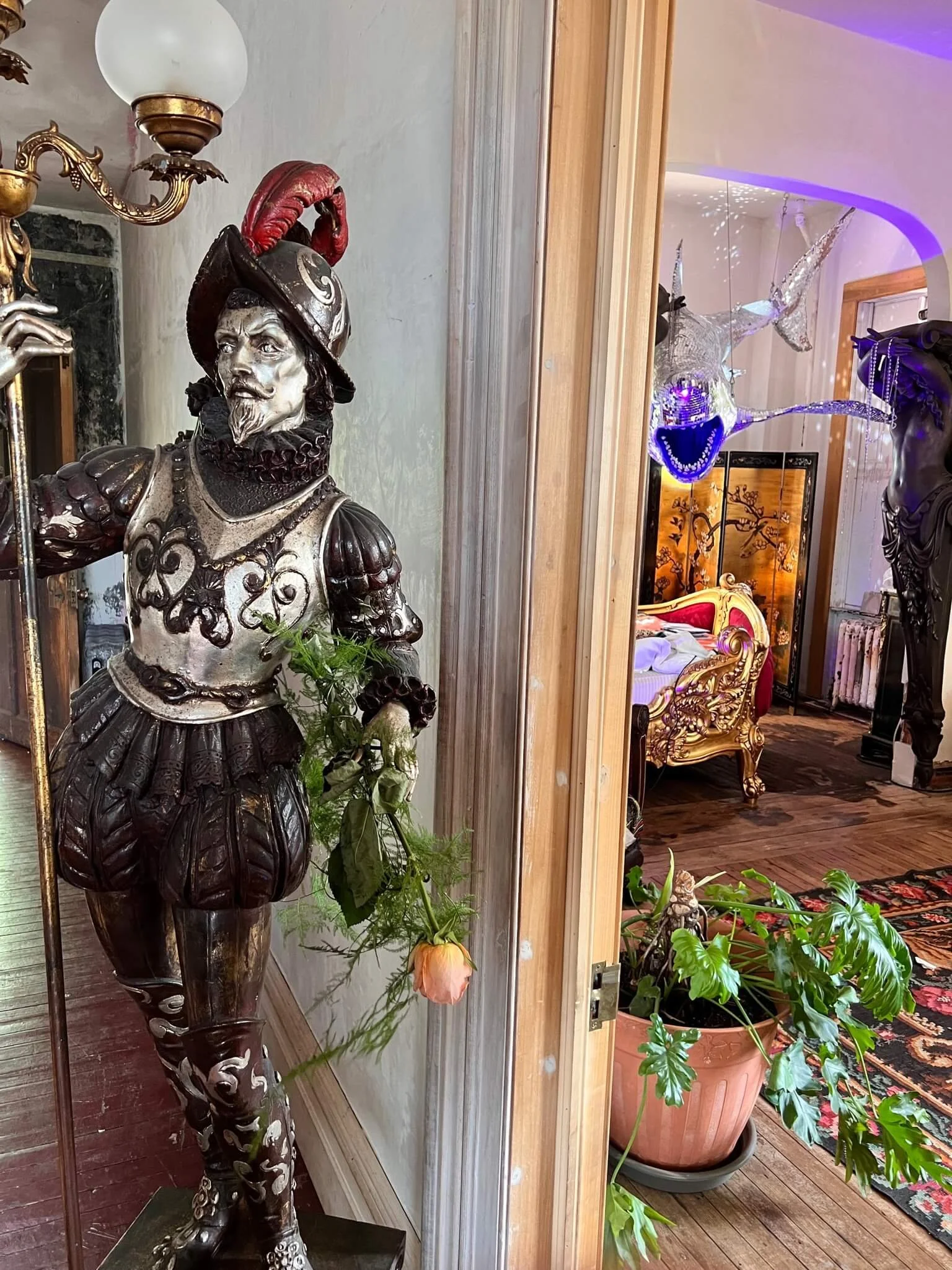 A decorative sculpture of a man in historical attire holding a staff, with a red feathered hat, standing indoors near a plant and a mirror.