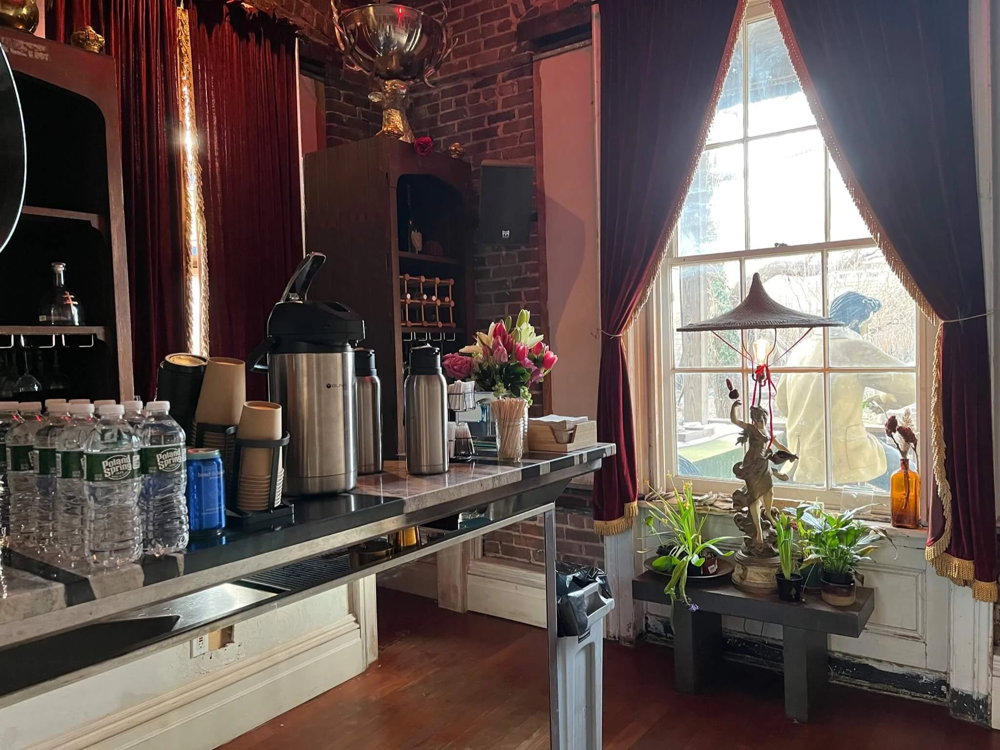 A cafe or restaurant interior with a counter on the left holding water bottles, cups, and coffee dispensers, and a large window with burgundy curtains on the right. Outside the window, a person wearing a yellow jacket is visible, and inside there is 