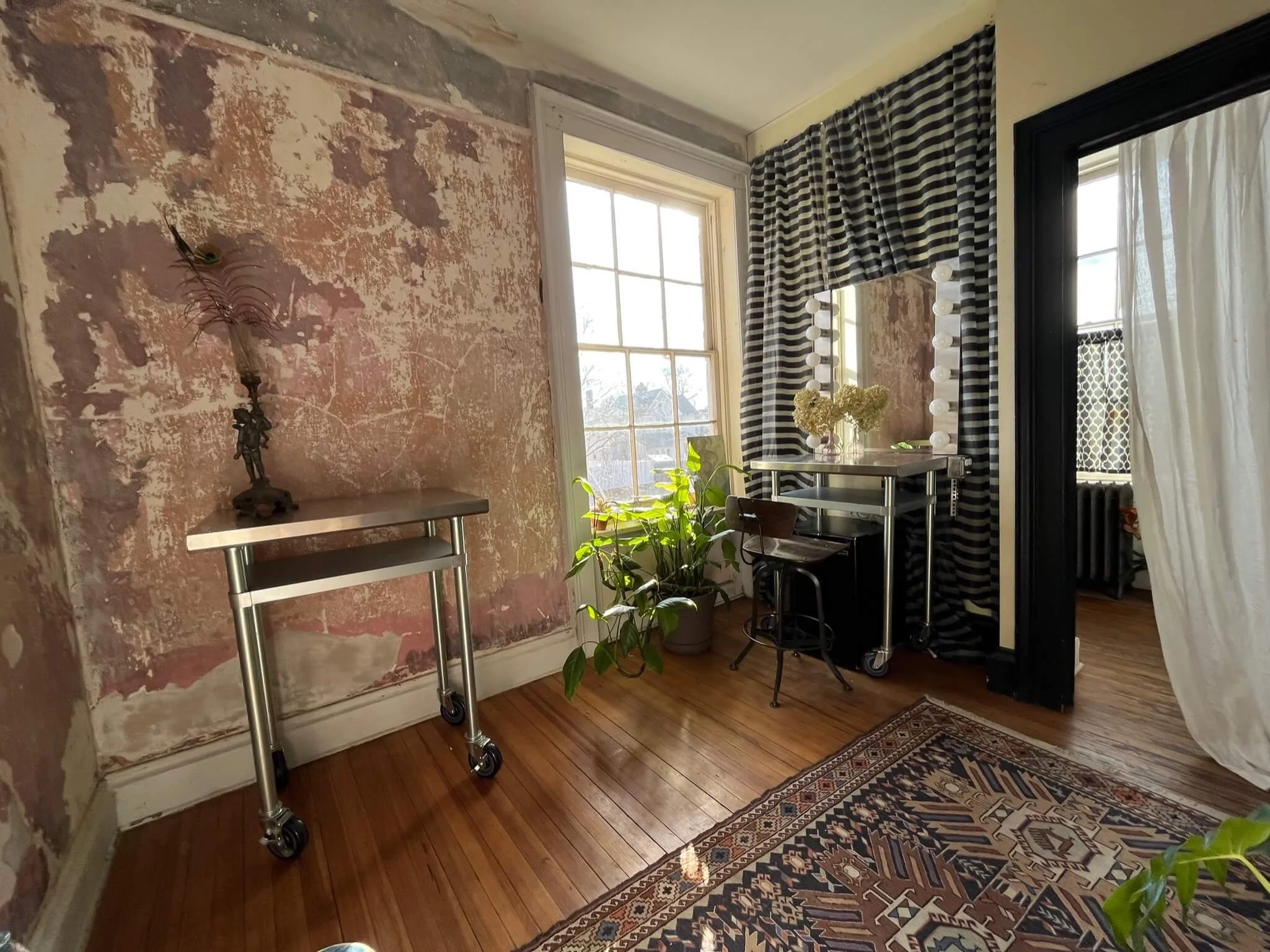 Interior view of a room with a distressed pink and beige wall, a wooden floor, and a window with white and black striped curtains. There's a small metal cart with a mirror and lights, a desk with flowers, a plant, and a patterned rug.