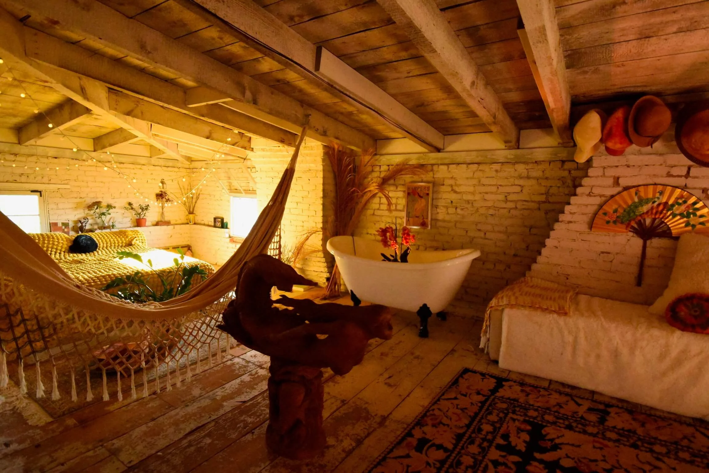 Cozy rustic living space with brick walls and wooden beams, featuring a hammock, a vintage clawfoot bathtub with plants, a couch with pillows and a decorative fan, and warm lighting.