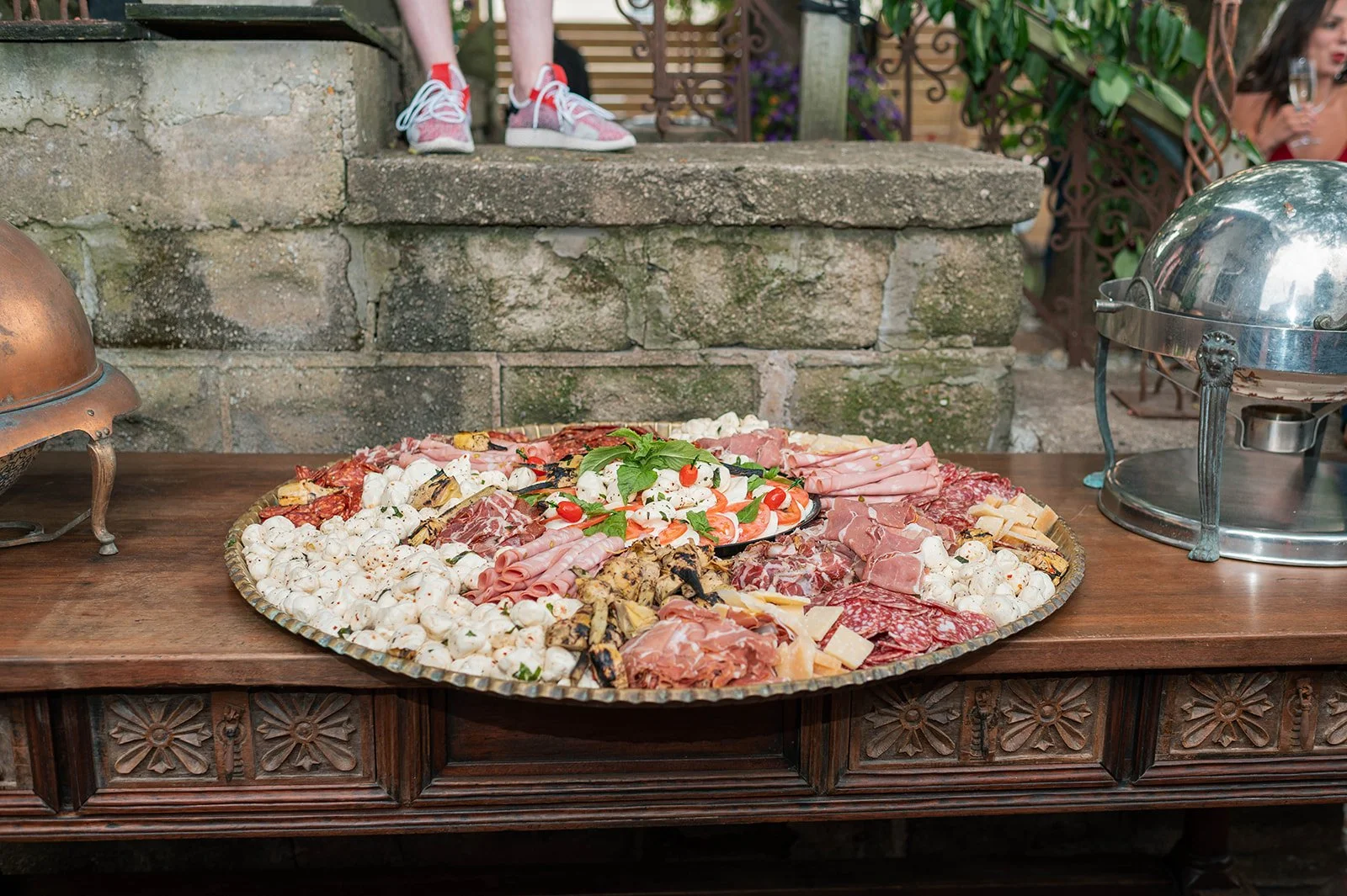 Large round platter of assorted meats and cheeses, garnished with basil and cherry tomatoes, on a wooden table outdoor, with a rustic stone wall in the background.