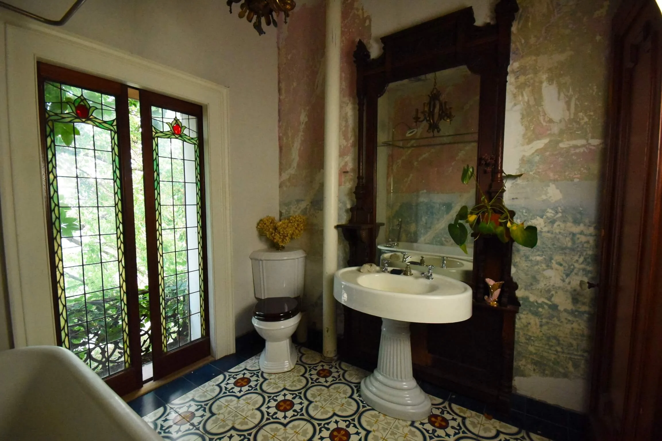 Vintage bathroom with stained glass door, ornate mirror, pedestal sink, toilet, and decorative patterned tile floor.