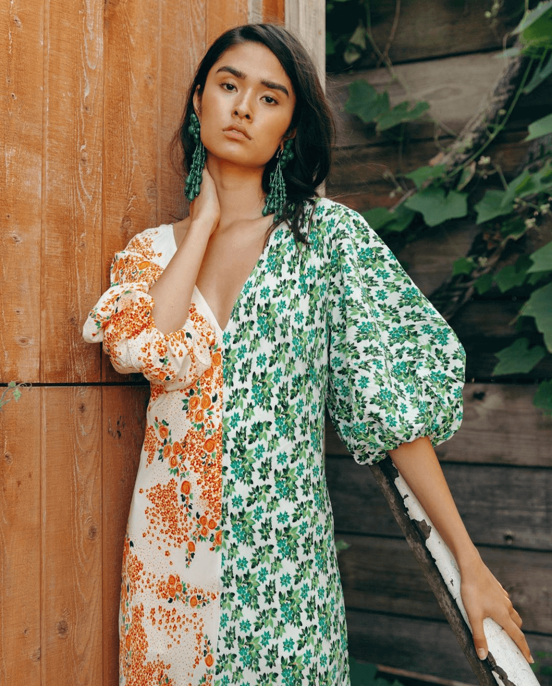 A young woman with dark hair and large green earrings wearing a floral print dress in green, white, and orange, standing outdoors near a wooden fence and green plants.