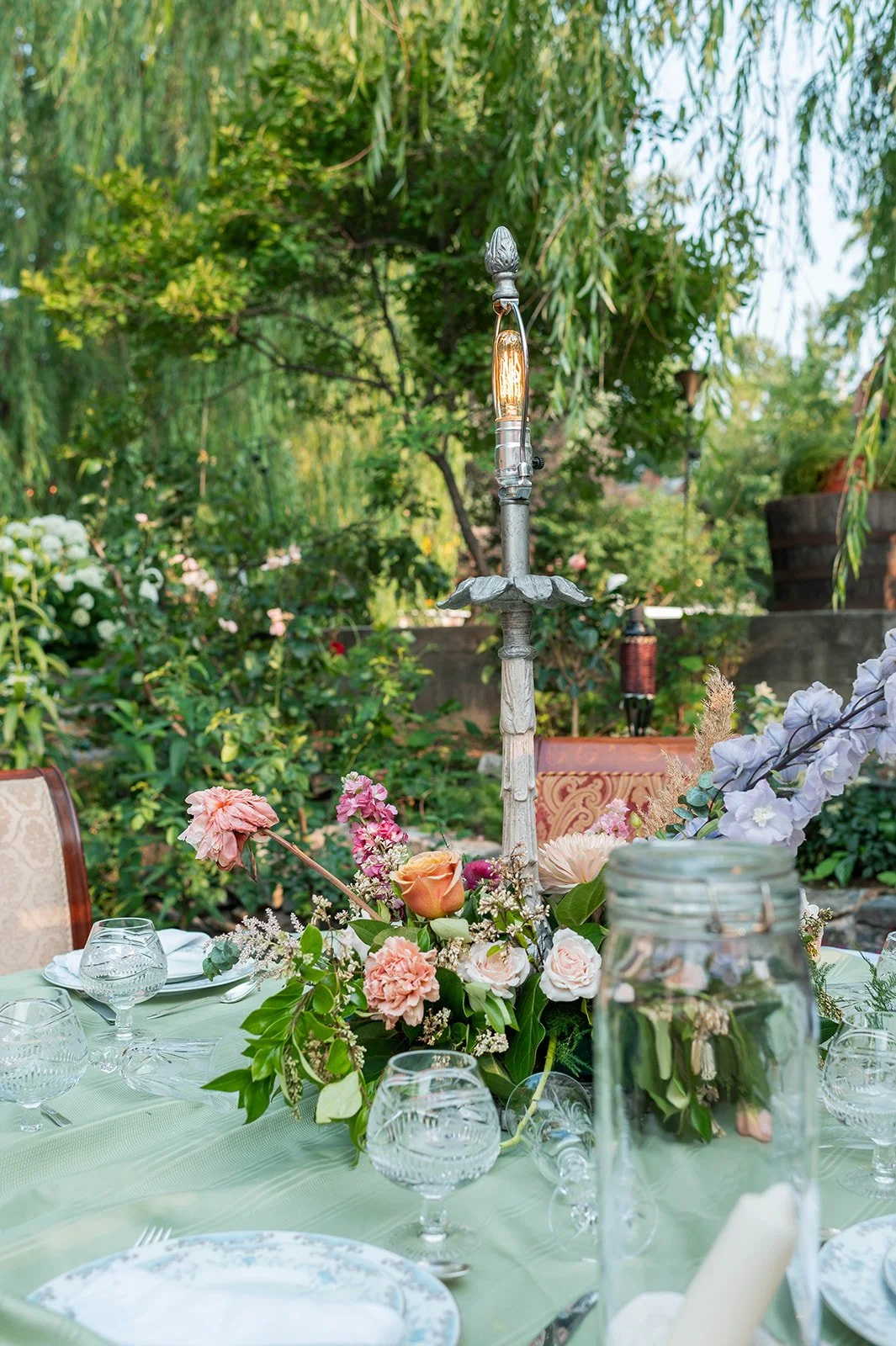 Table set for outdoor event with floral centerpiece, glassware, and a vintage-style lamp, surrounded by greenery.