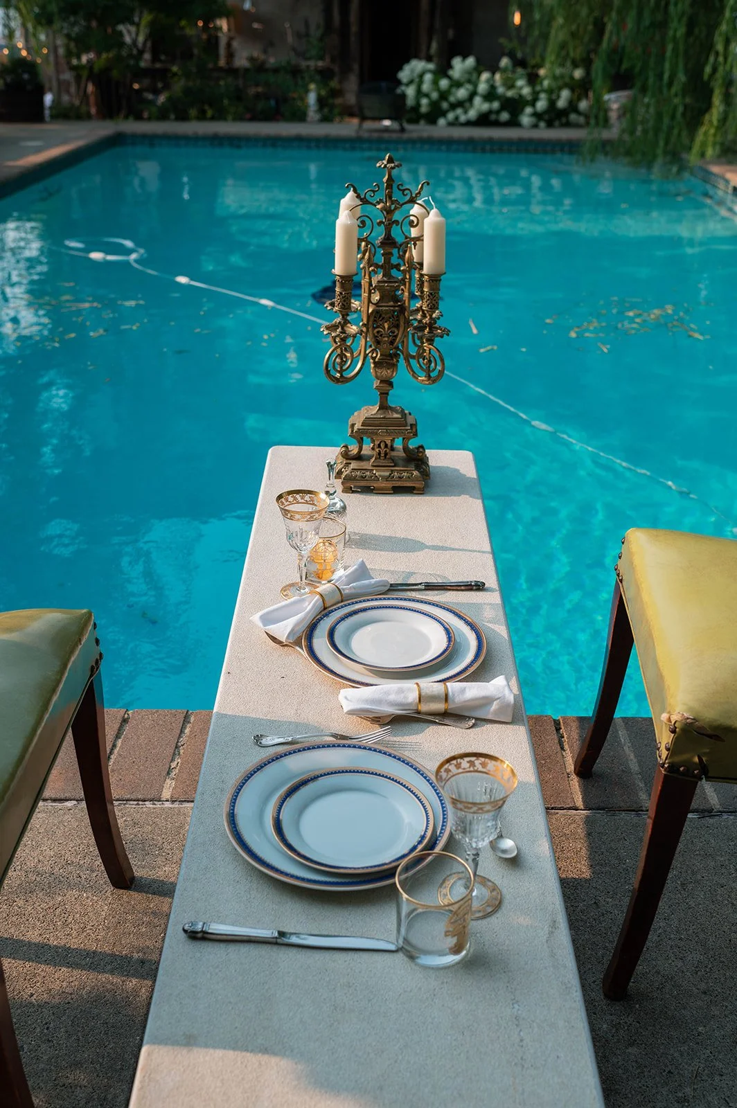 A dining setup with three plates, glasses, a napkin, and silverware on a table next to a swimming pool. A vintage chandelier is placed on the table, and the pool is outdoors surrounded by greenery.
