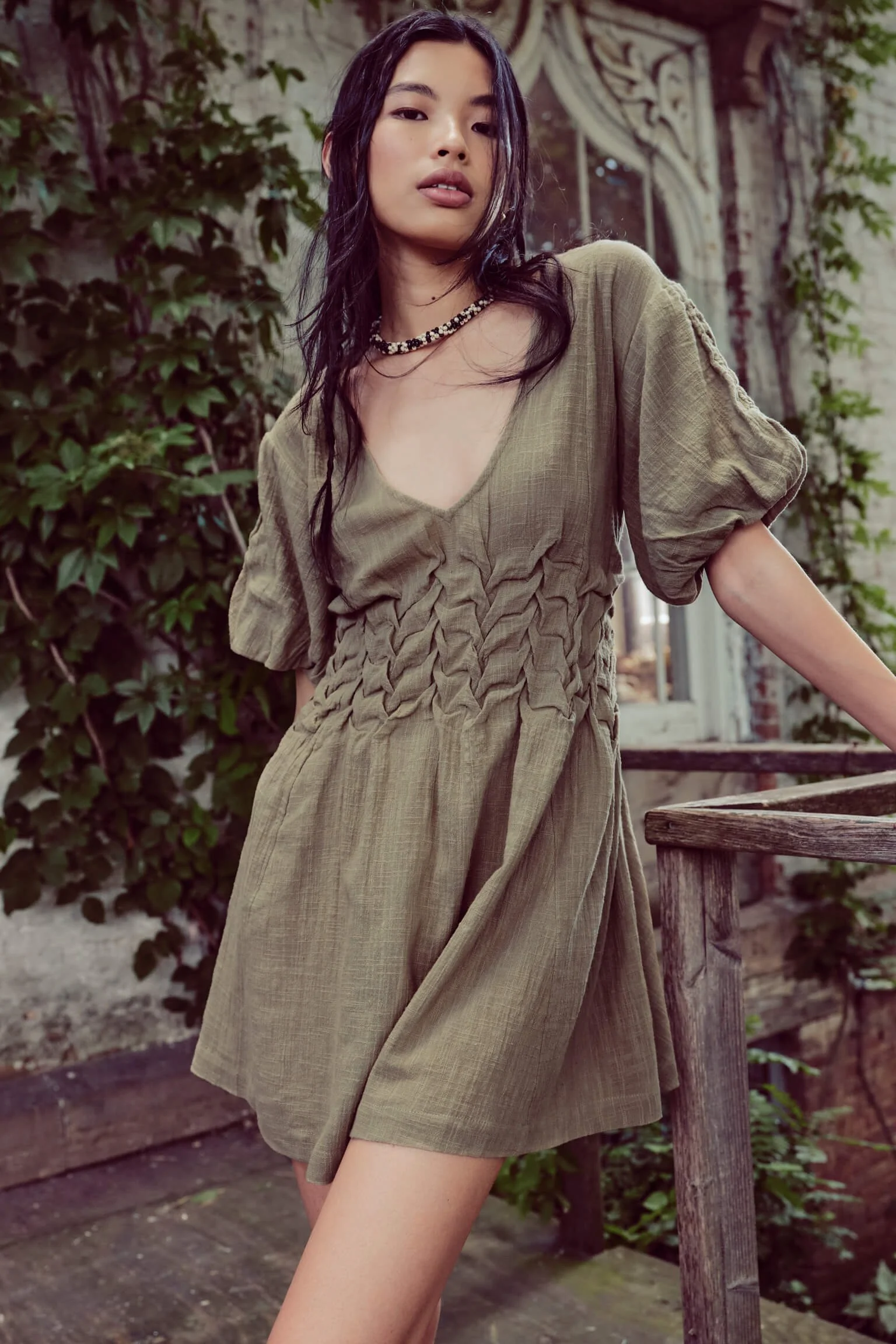 A young woman with long dark hair poses outdoors, wearing a khaki-colored dress with puffed sleeves and gathered details, and a beaded necklace.