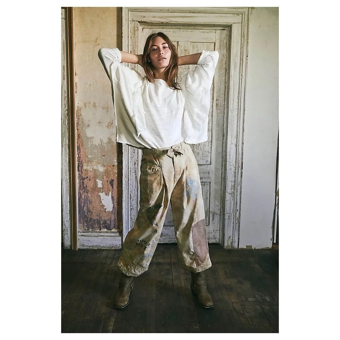 A young woman with brown hair posing in front of an old, distressed wooden door. She is wearing a loose, white long-sleeve top, baggy paint-stained beige pants, and brown boots, with her hands resting behind her head.
