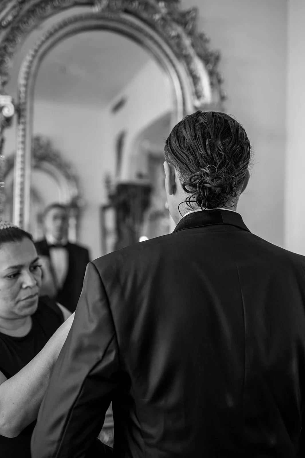 A person getting their hair styled in front of a mirror with ornate frames, with another person assisting, and a third person visible in the background also looking into a mirror.