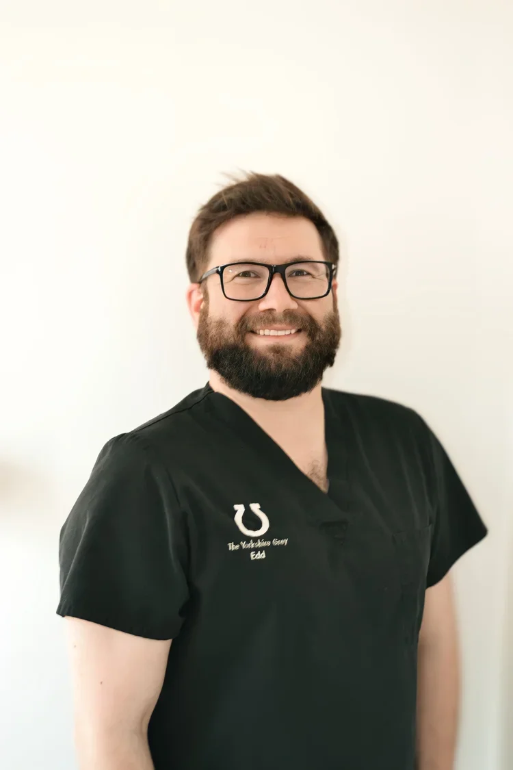 Portrait of a smiling man with glasses and a beard wearing a black medical scrub top against a light background.