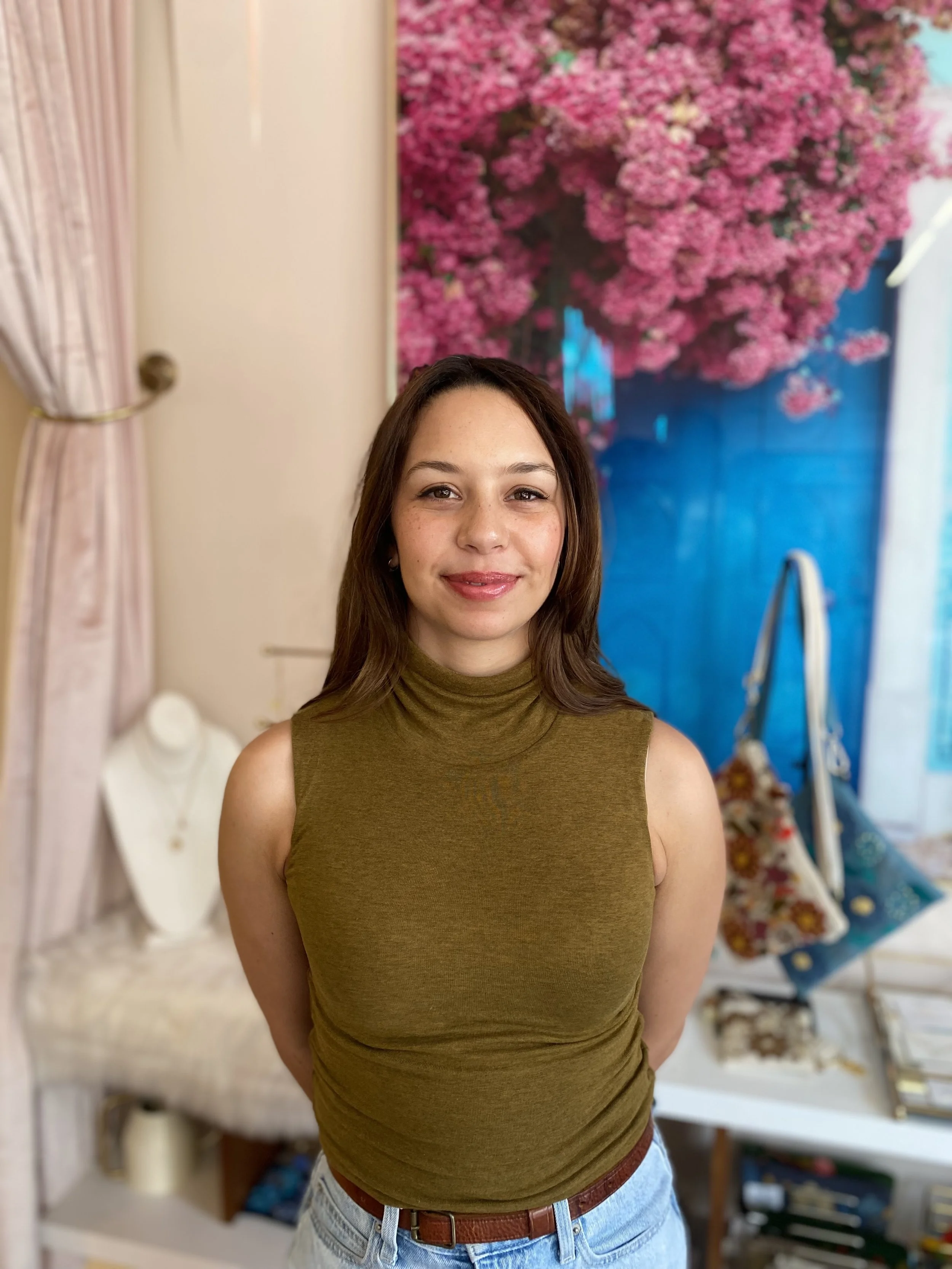 Woman in a green sleeveless top standing in a boutique with floral decorations and purses in the background.