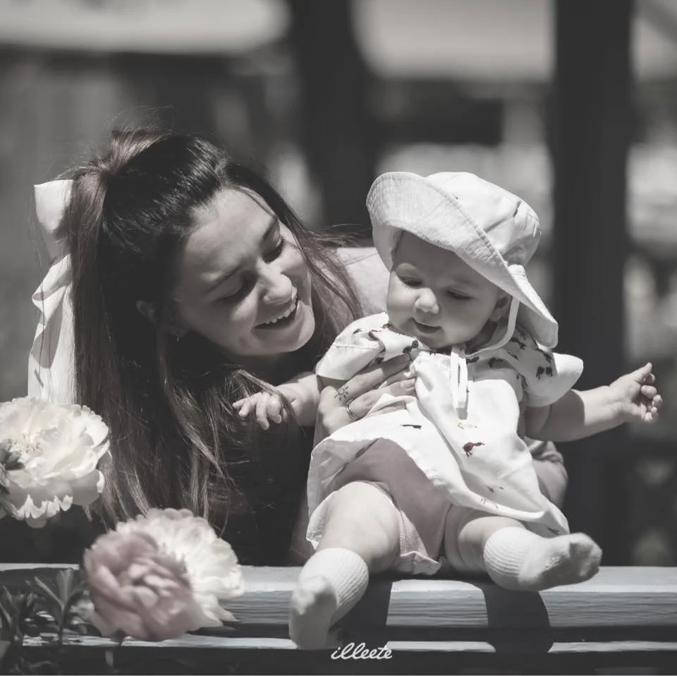 A woman smiling and holding a baby girl who is sitting on a wooden surface with flowers nearby. The baby wears a sun hat and socks, and both are outdoors.