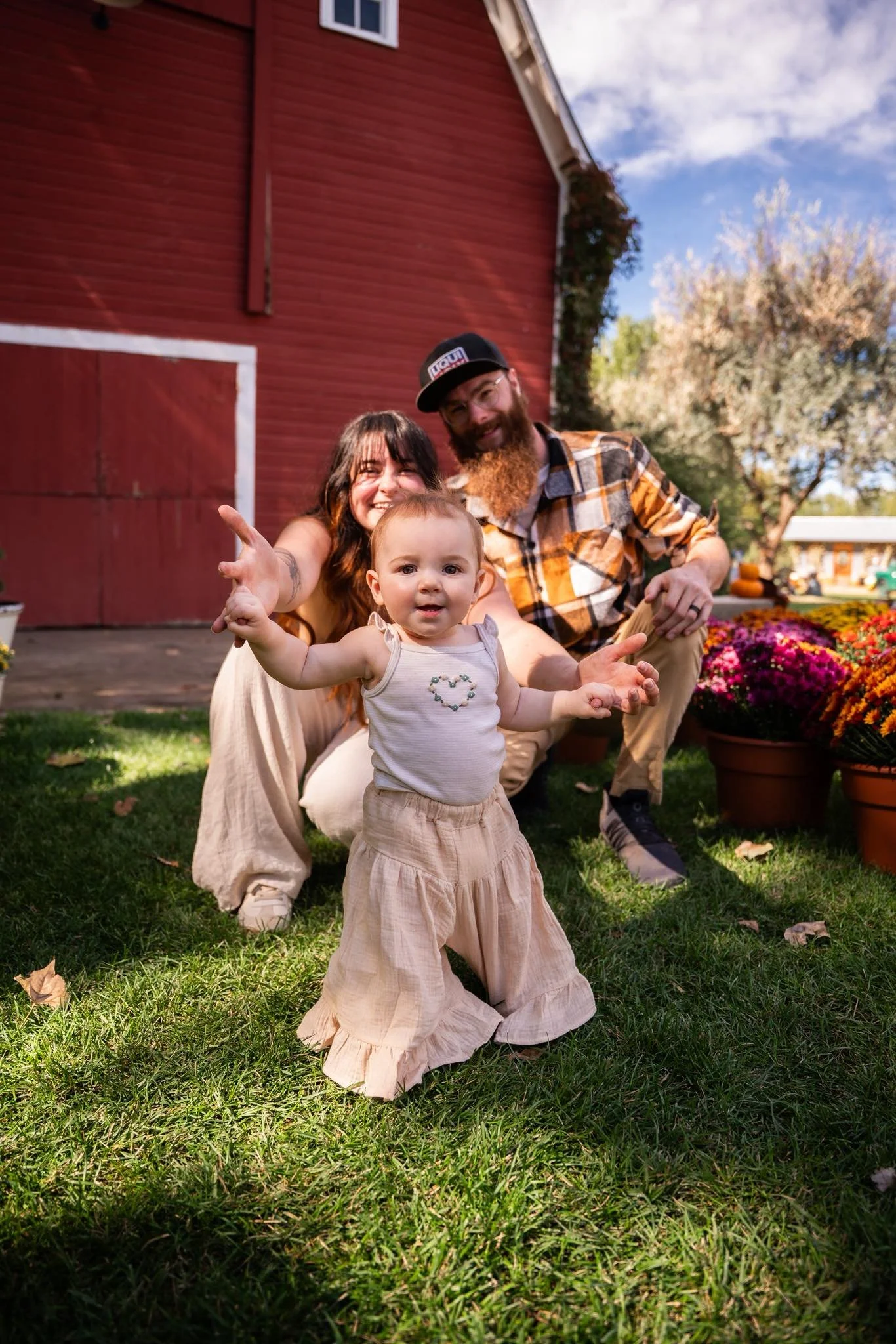 A family of three outdoors on a sunny day, with a red barn and potted flower plants in the background. The baby girl is standing on the grass, with the parents behind her.
