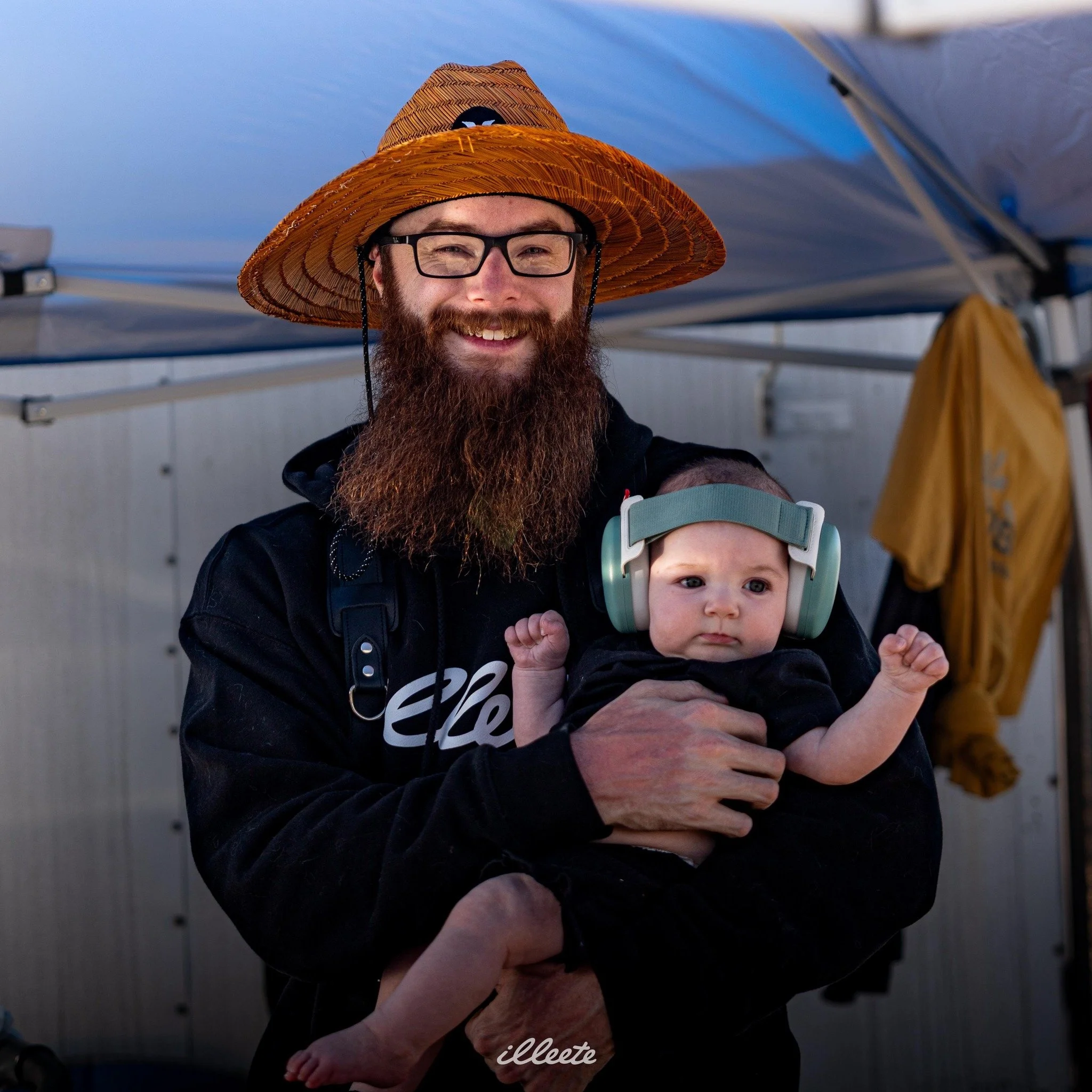 A man with glasses and a large beard wearing a straw hat is holding a baby with headphones. They are outdoors under a tent.