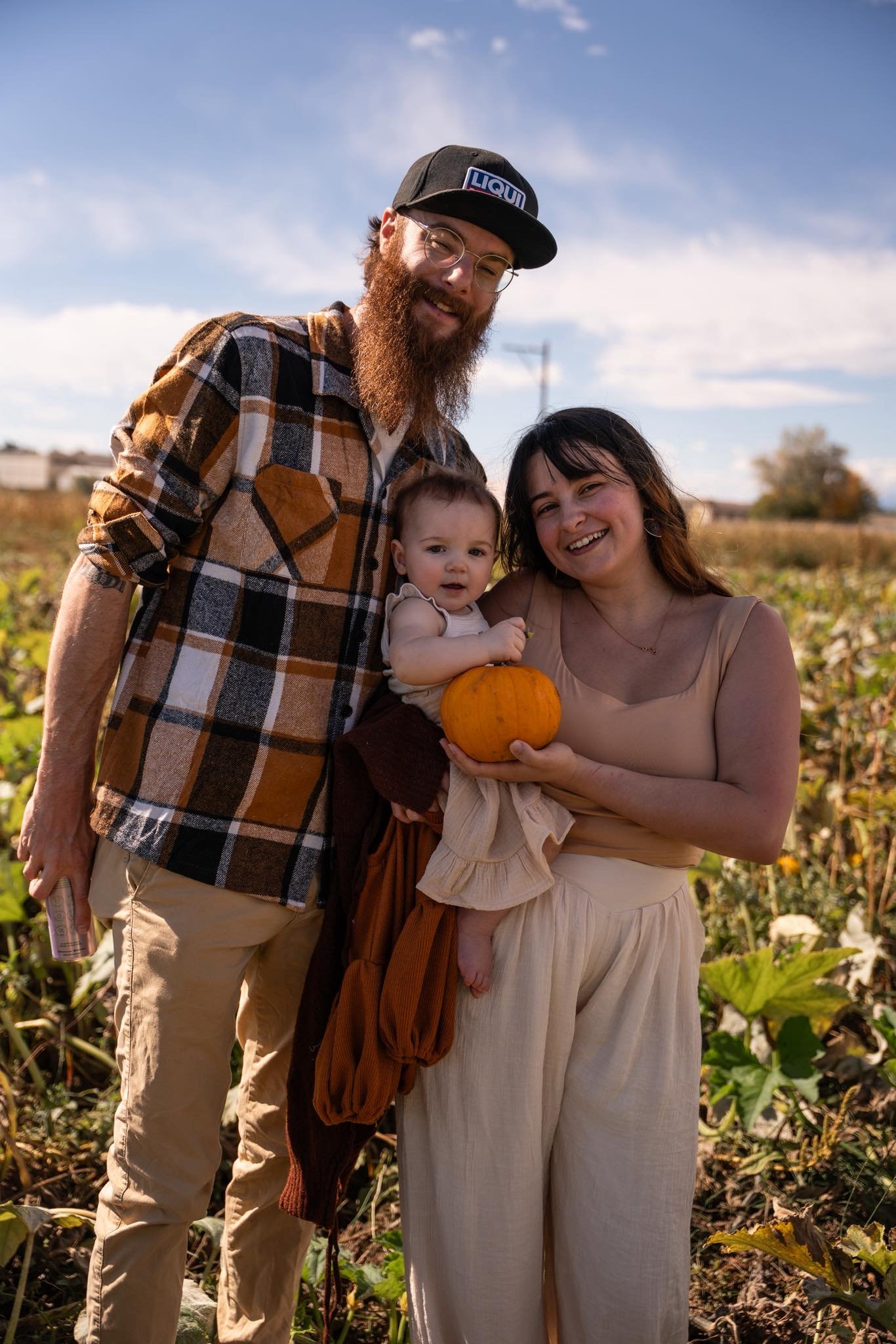 A family of three, including a man with a beard and glasses, a woman, and a young girl holding a pumpkin, standing outdoors in a pumpkin patch with a blue sky and clouds.