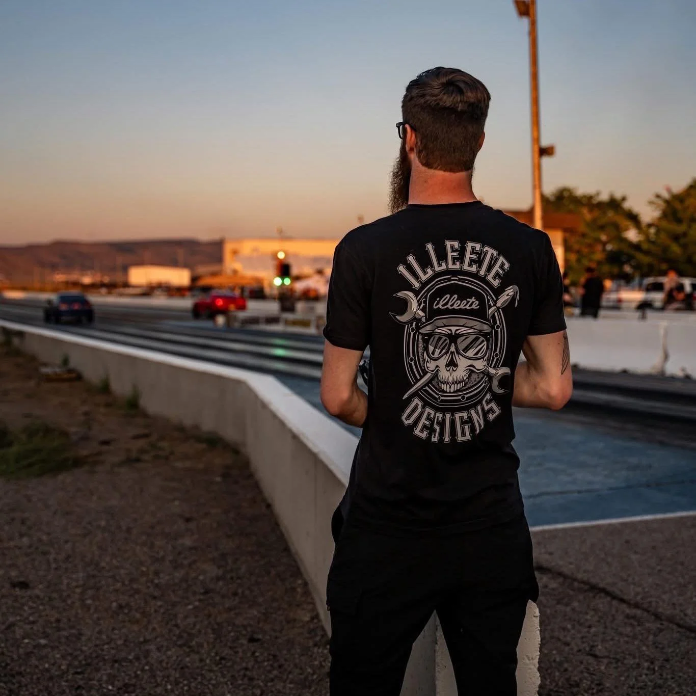 A man with a beard and glasses standing on the side of a highway at sunset, wearing a black T-shirt with a skull graphic and 'Illteet Designs' logo on the back.