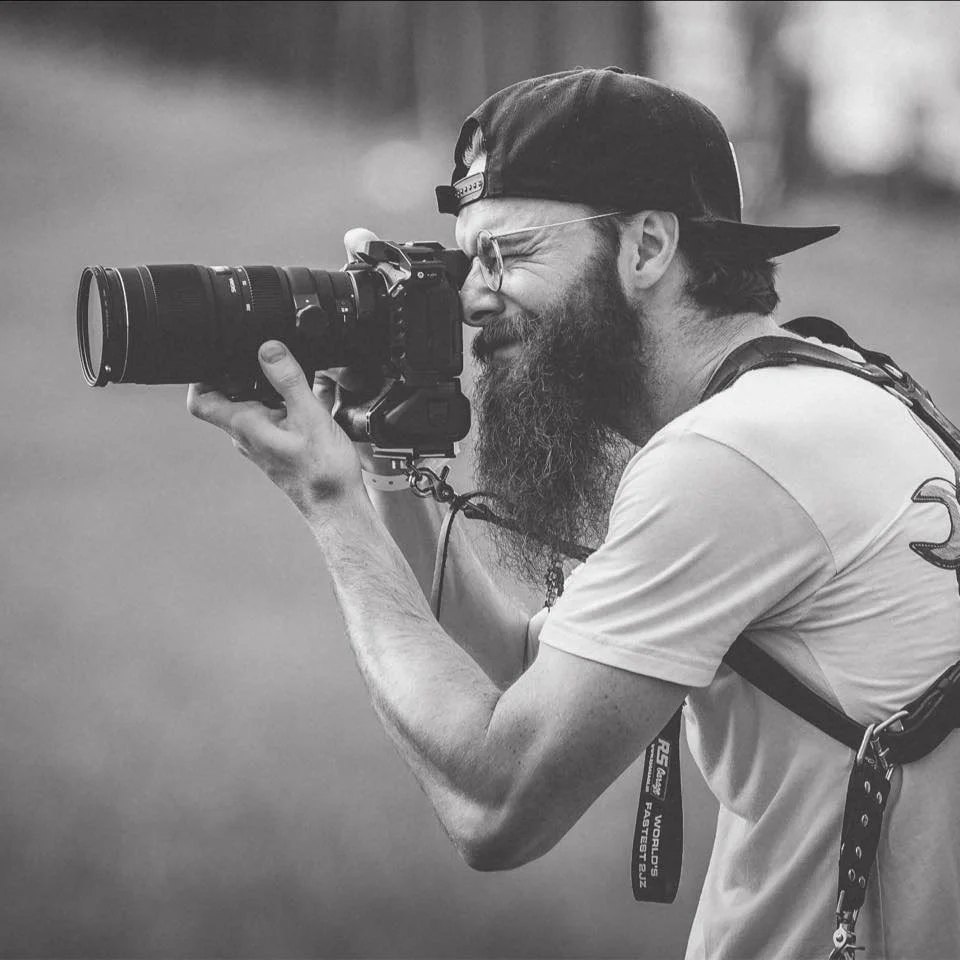 A man with a beard, glasses, and wearing a backwards cap is holding a camera and taking a picture. He seems focused and is looking through the camera viewfinder.
