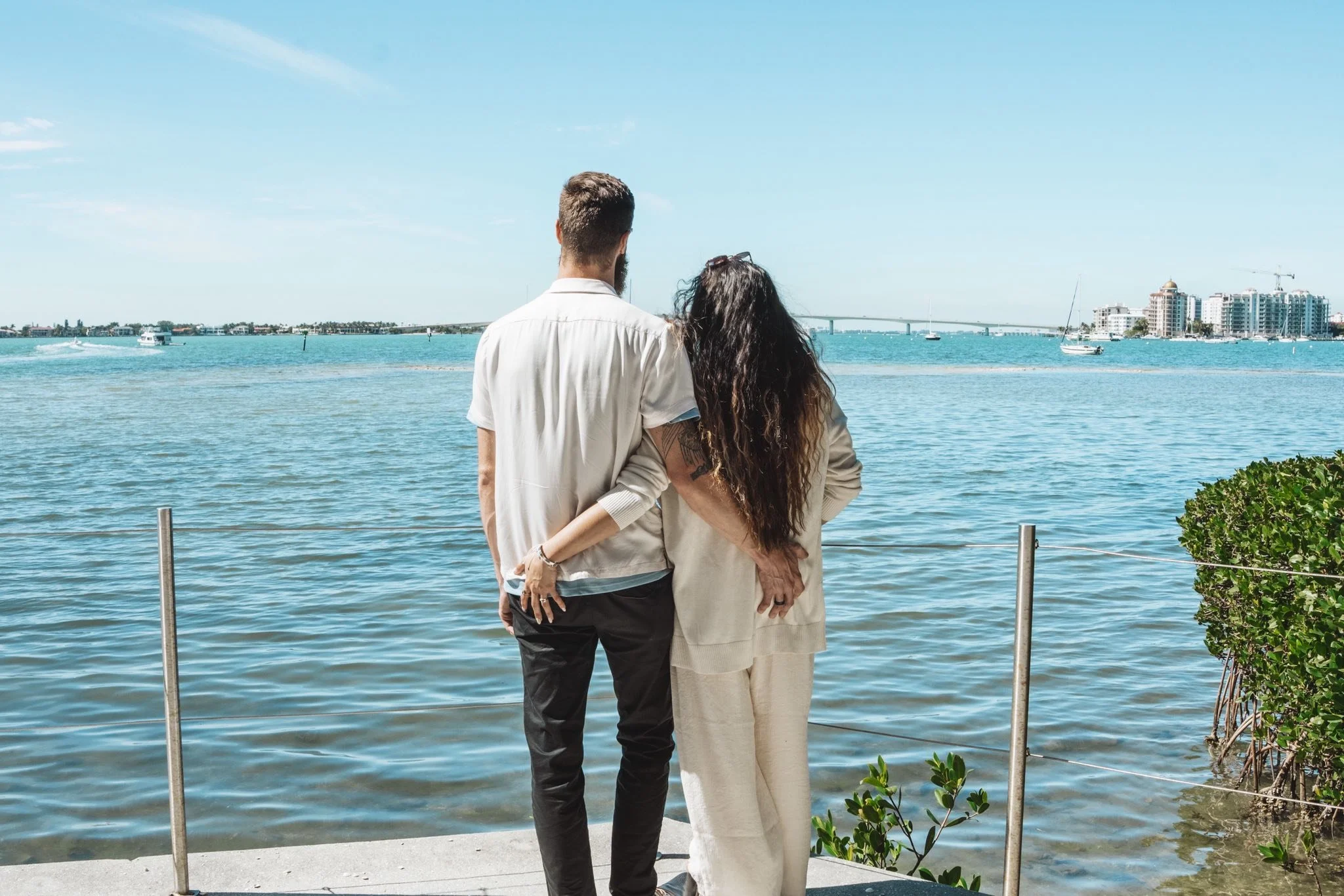 A couple standing by a waterfront, facing the water and taking in the view, with city buildings and a bridge in the background.