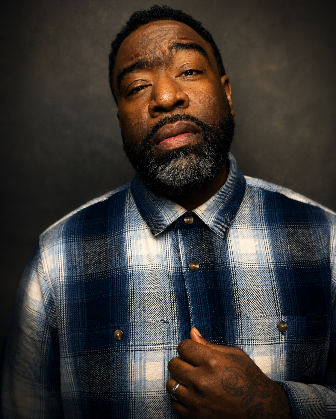 A man with a dark beard and mustache wearing a blue and white plaid shirt, looking directly at the camera against a dark background.