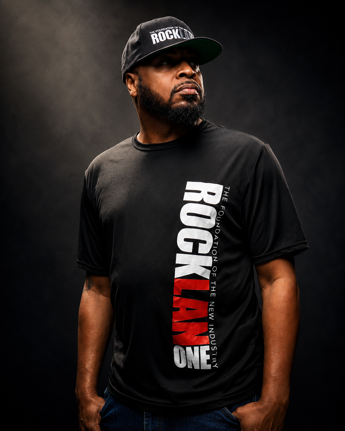 A man wearing a black cap and black T-shirt with the words 'The Foundation of the Rockland' and 'One' in bold letters, standing against a dark background.
