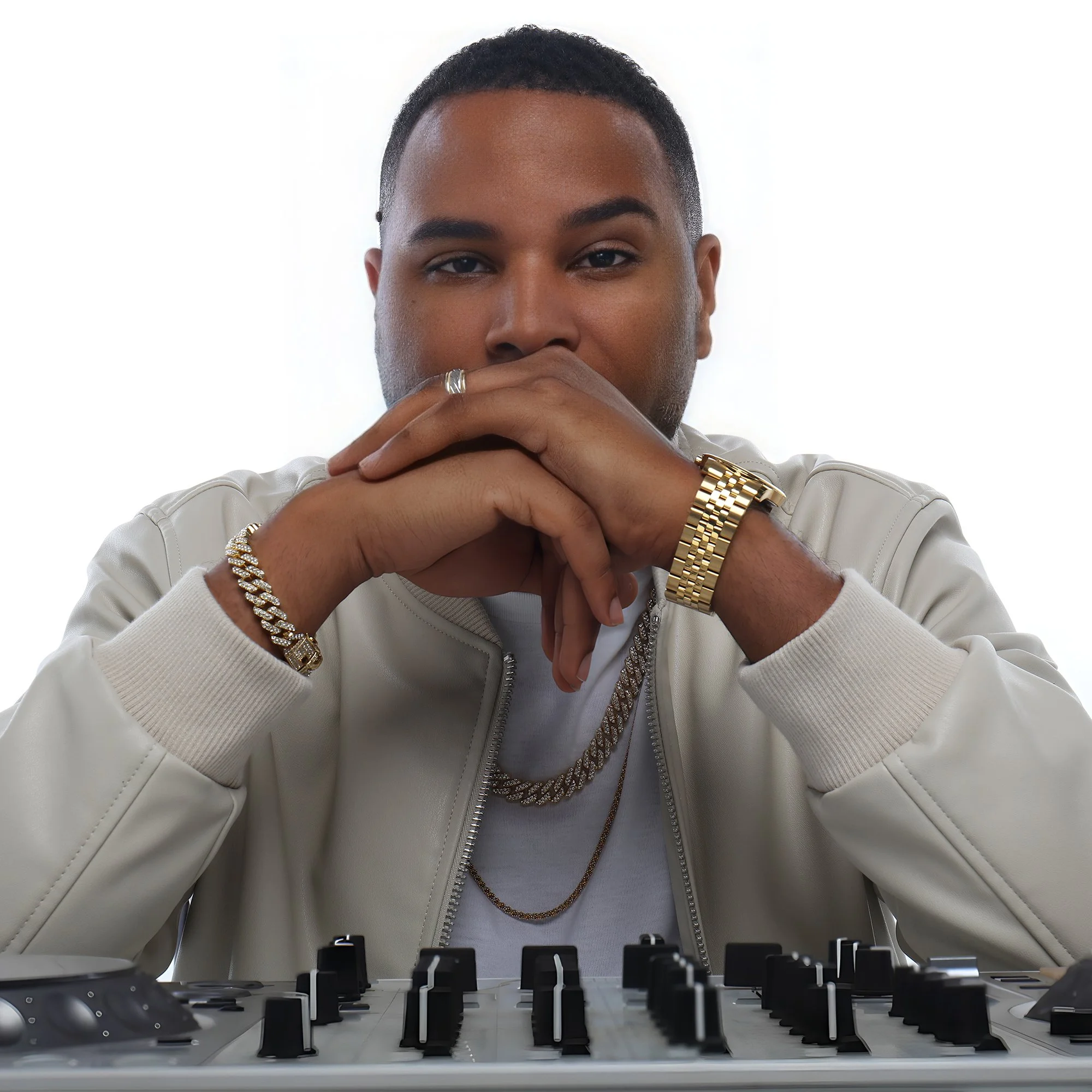 A young man with short black hair, wearing a white jacket, gold jewelry including a watch, bracelet, chains, and a ring, sitting behind a music mixer or synthesizer with his hands clasped in front of his face, gazing intently at the camera.