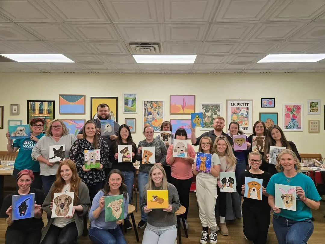 Group of people holding painted dog portraits in an art gallery or classroom.