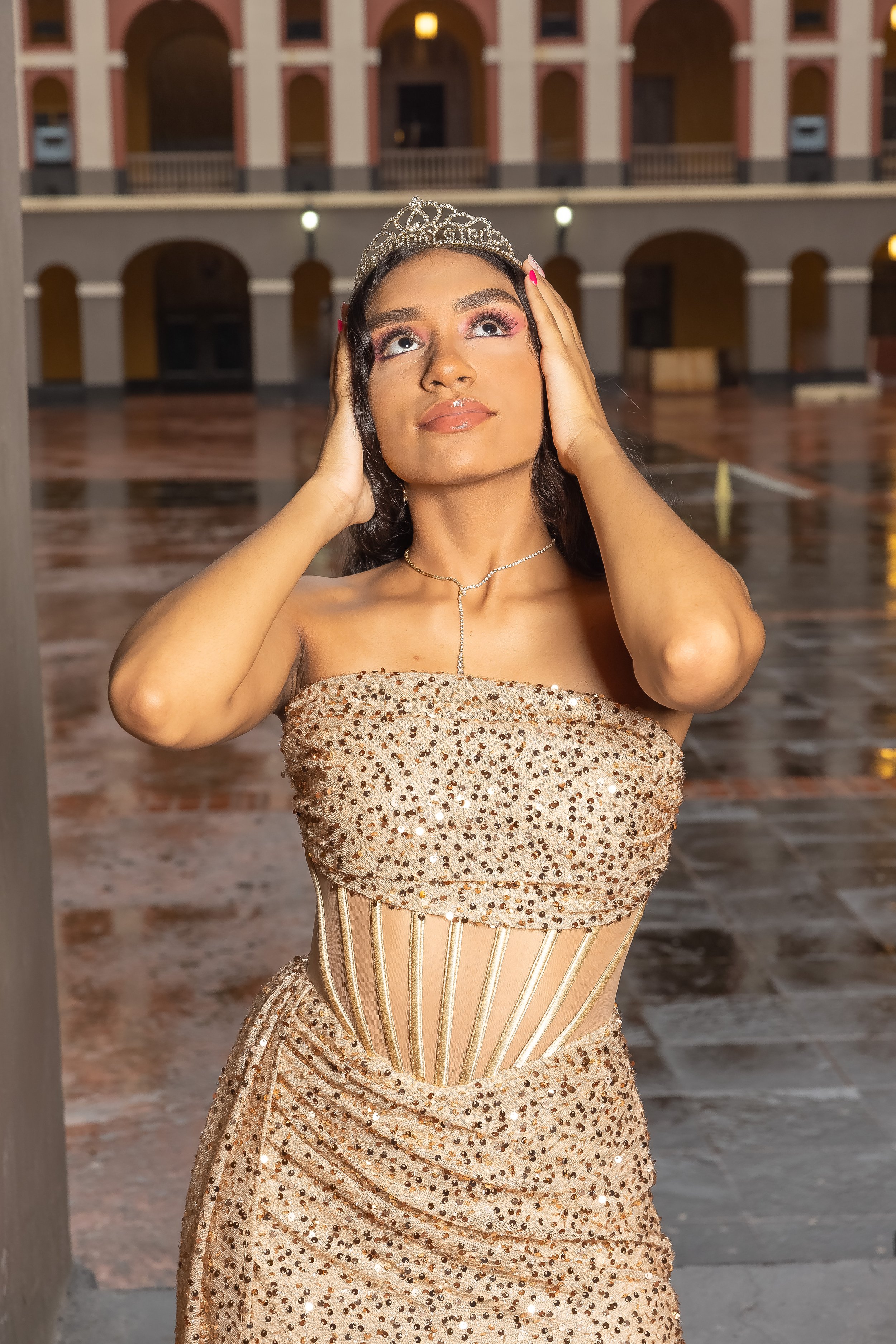 Joven con vestido dorado y tiara, en un patio con arcos, ajustándose el cabello y luciendo feliz.