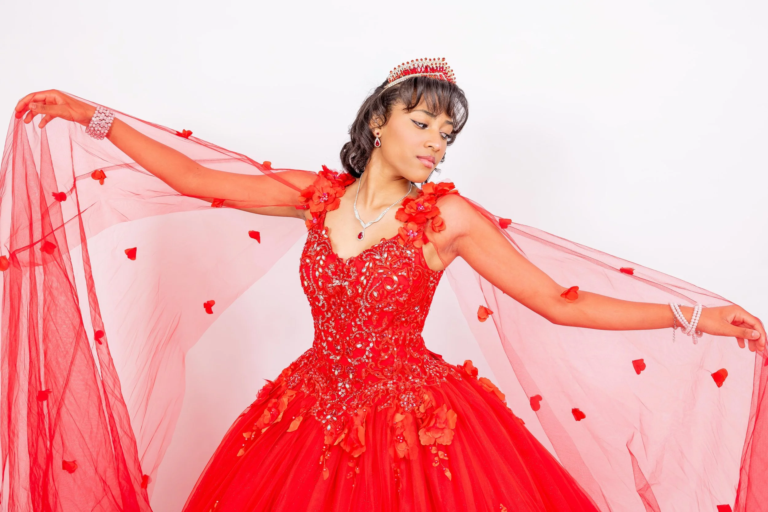 Mujer vestida con un elegante vestido rojo con detalles de pedrería, coronada y usando joyería, posa con una tela transparente decorada con pétalos rojos contra un fondo blanco.
