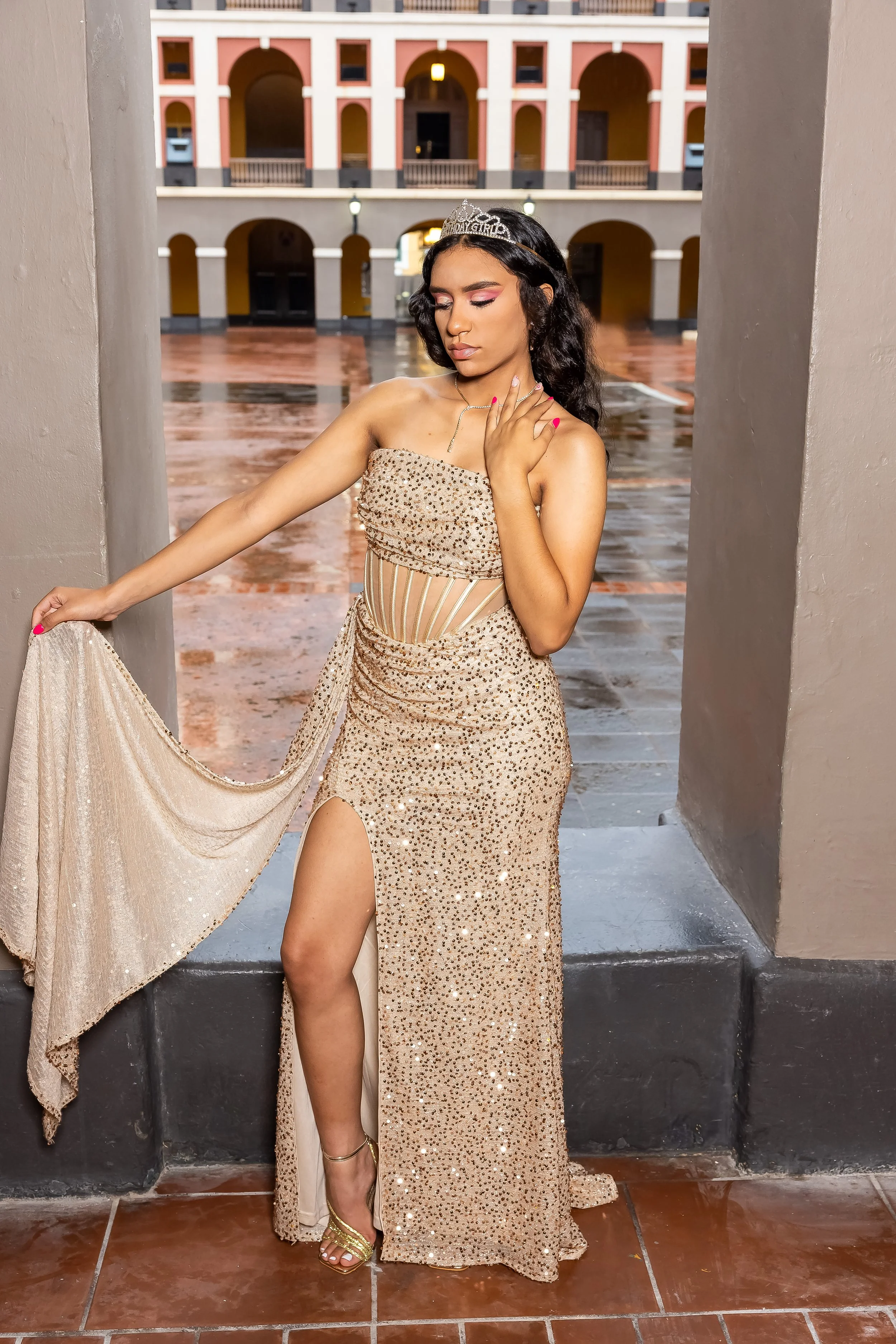 Joven mujer vestida con vestido de noche dorado, con tiara, posando en un fondo de arquitectura colorida y suelo de tierra mojada.