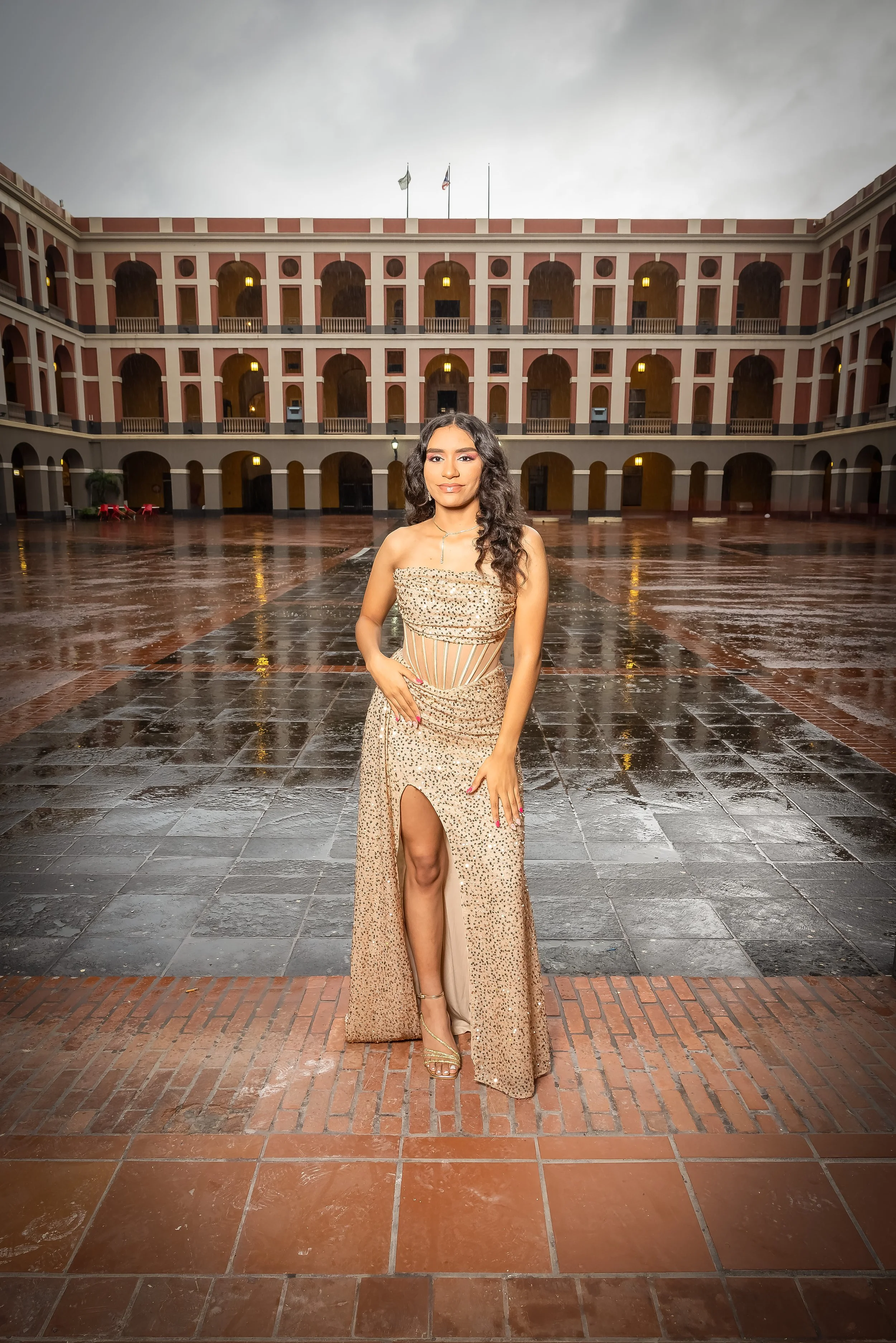 Una joven vestida con un vestido dorado brillante de noche, posando en un patio interior de un edificio colonial con múltiples arcos y banderas en el techo, tras una lluvia.