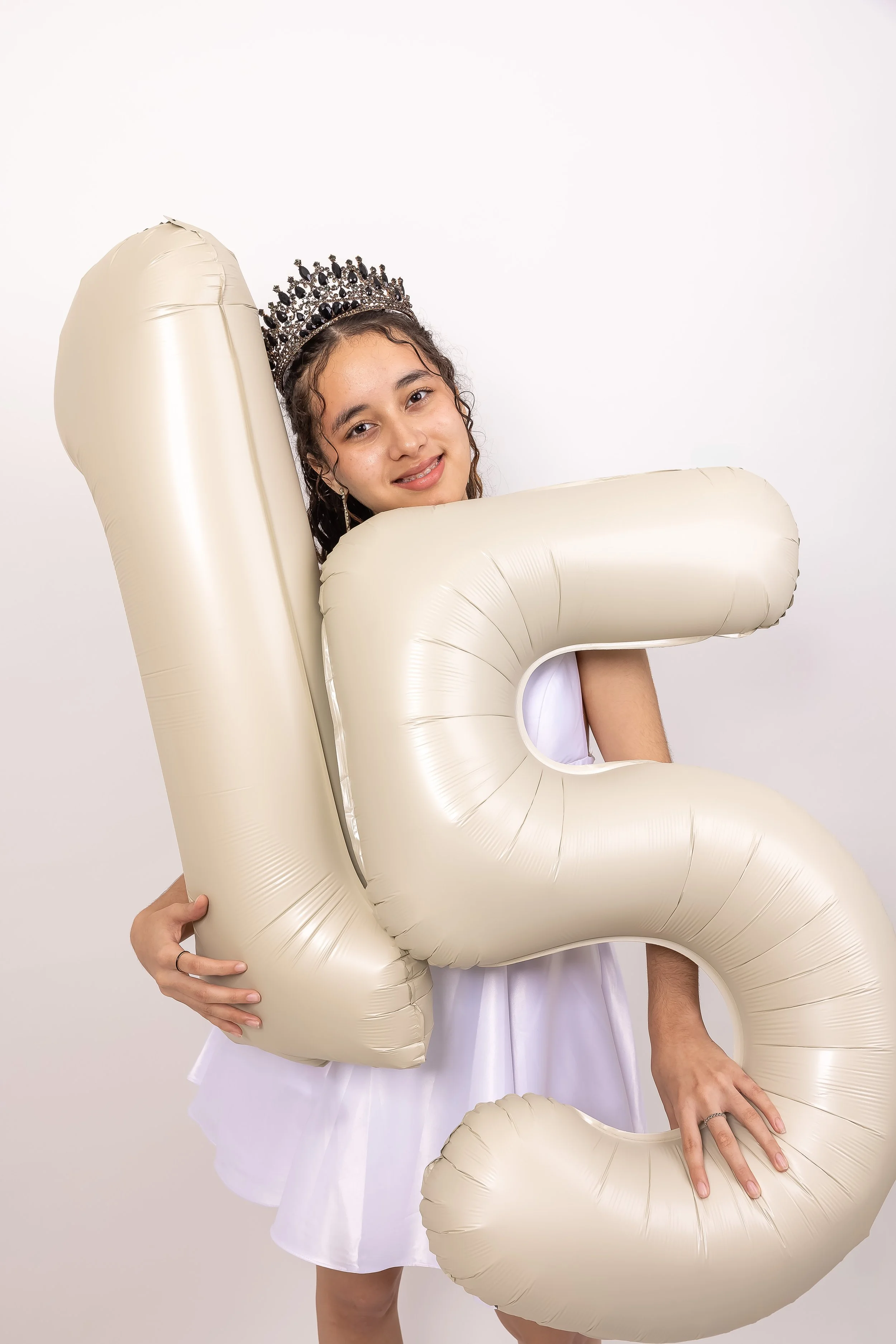 Juventud con corona y vestido blanco sostiene globos en forma de números 1 y 2, celebrando un cumpleañ o.