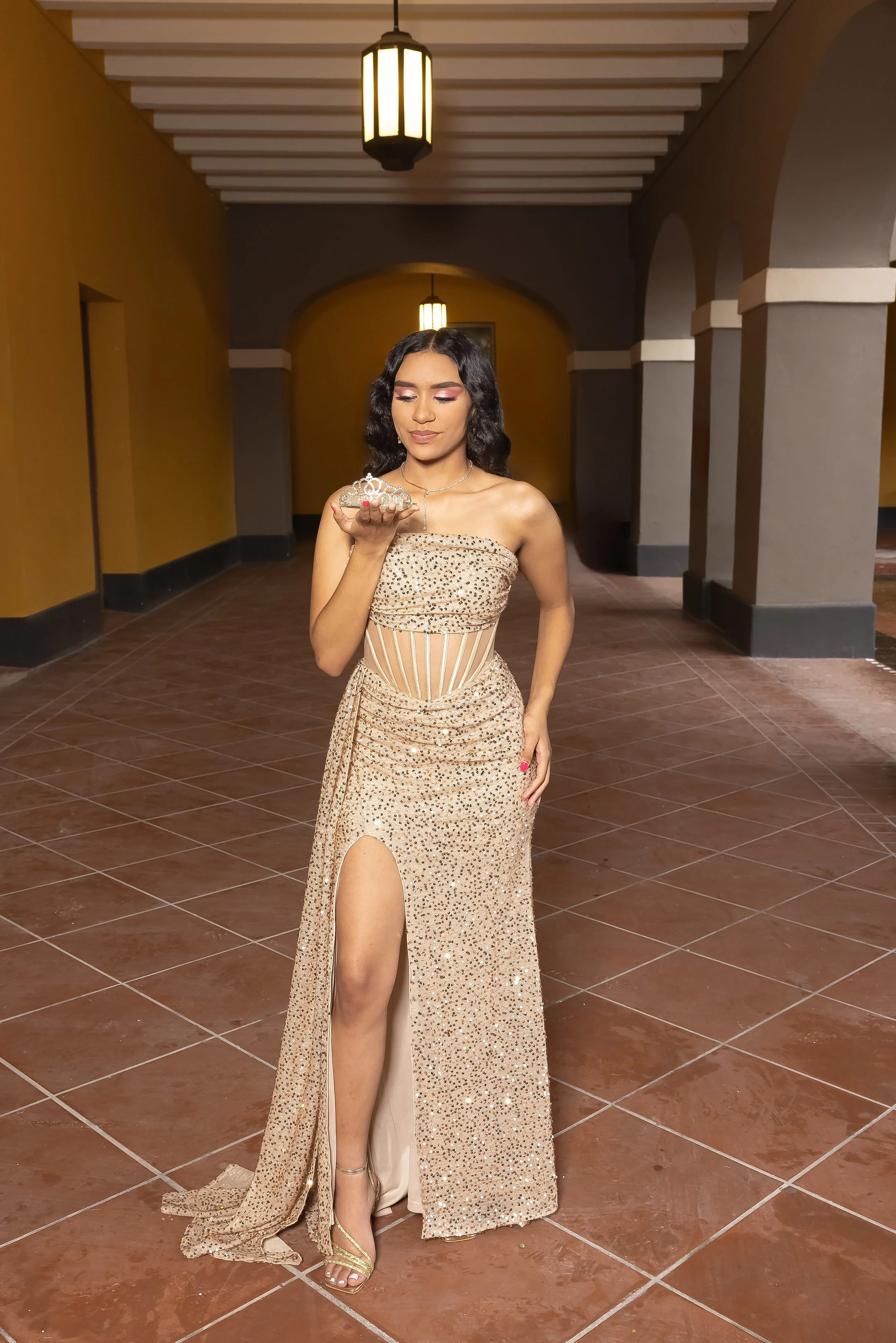 Joven mujer en vestido de noche dorado con abertura en la pierna, mirando un arete en su mano en un pasillo con luz cálida y paredes de color mostaza y gris.