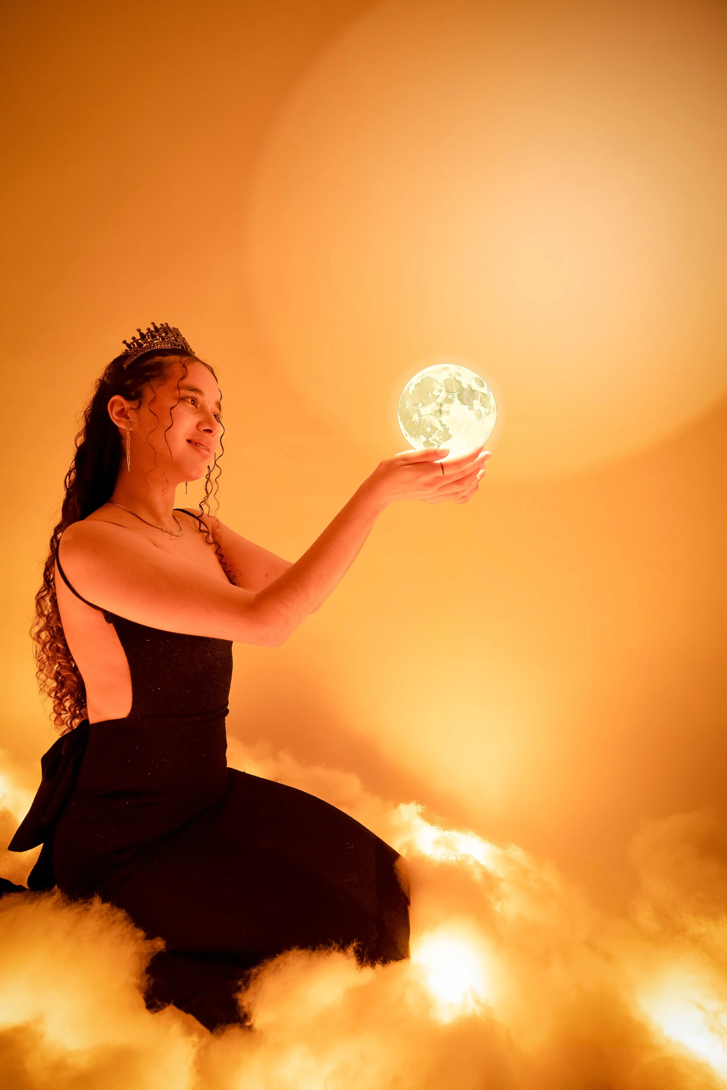 Mujer con diadema y vestido negro sentado entre nubes doradas, sosteniendo una luna llena en sus manos, con un fondo de cielo naranja.