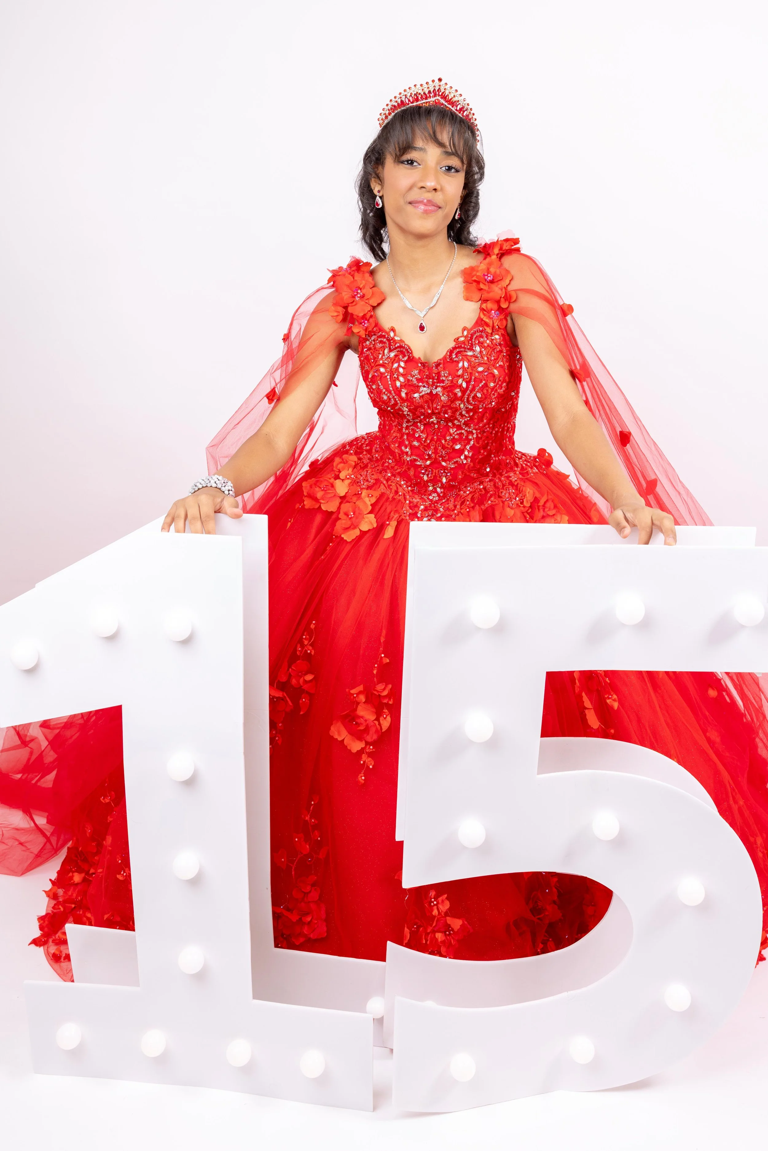 Mujer en vestido largo de color rojo con corona y collar, sosteniendo letras blancas con luces y un número 5, en fondo blanco.