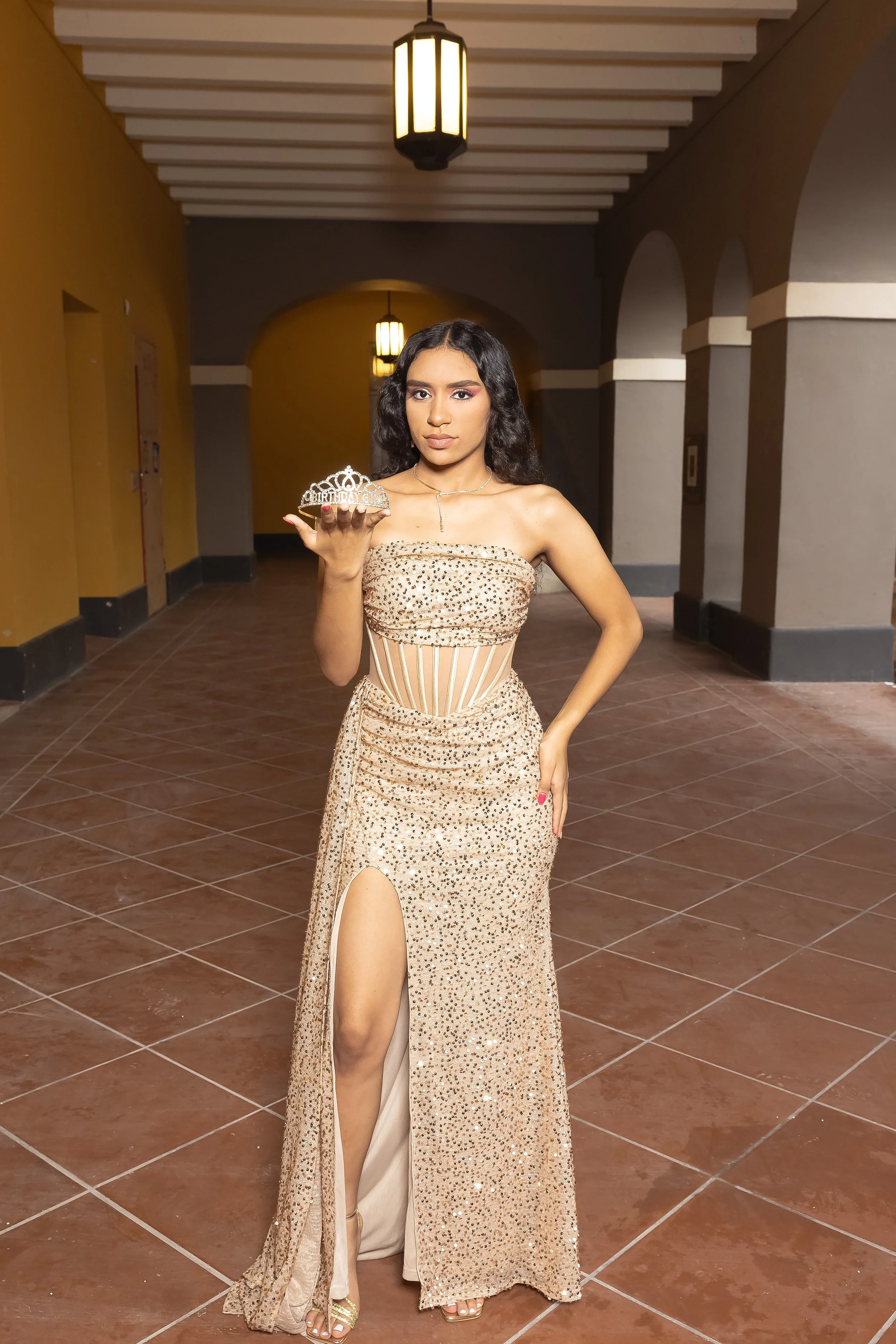 Joven mujer en vestido de gala dorado con abertura en la pierna, sosteniendo una tiara de cumpleaños en un pasillo con techos decorativos y faroles. 