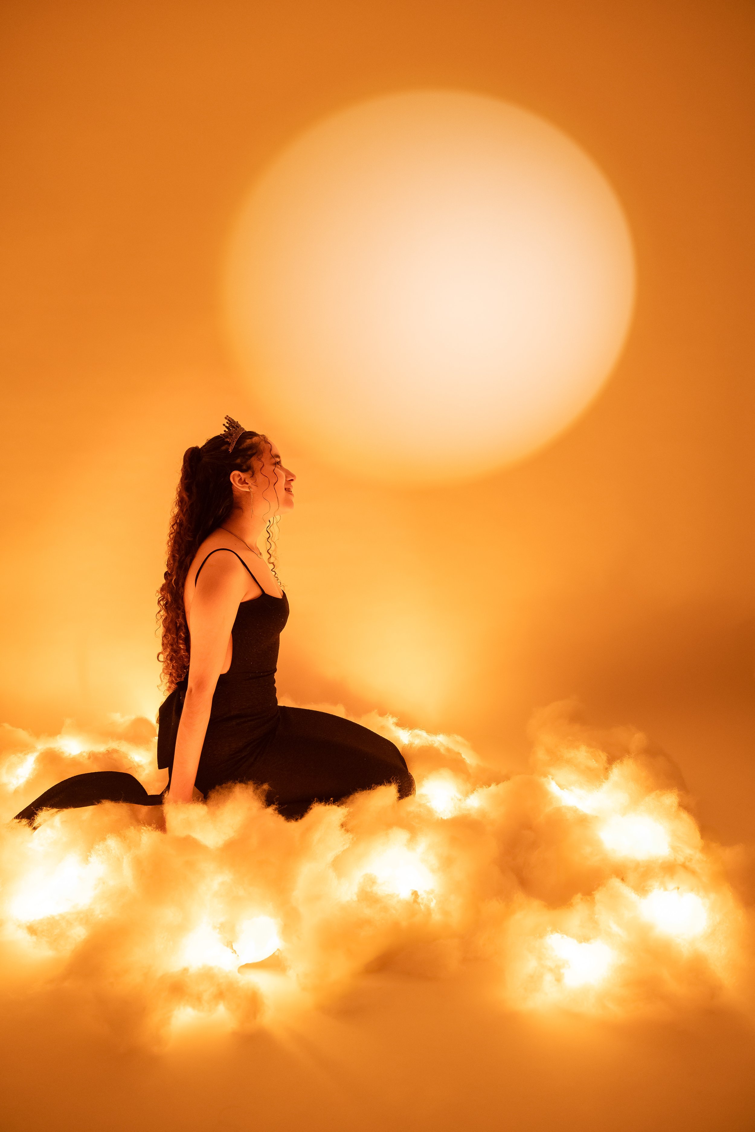 Una mujer con corona de princesa, vestida de negro, sentada sobre nubes iluminadas con luz cálida, con un gran sol brillante en el cielo naranja de fondo.