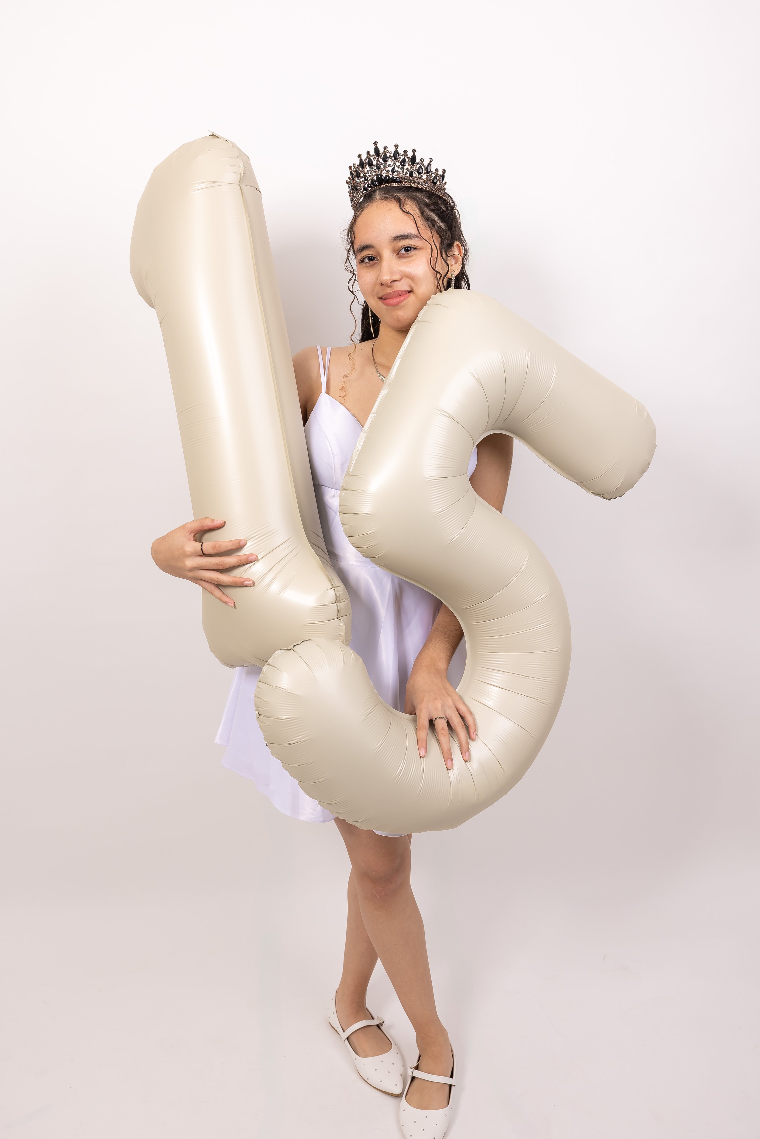 Joven con corona y vestido blanco sosteniendo globos en forma de número 1 y 3, celebrando un cumpleaños.