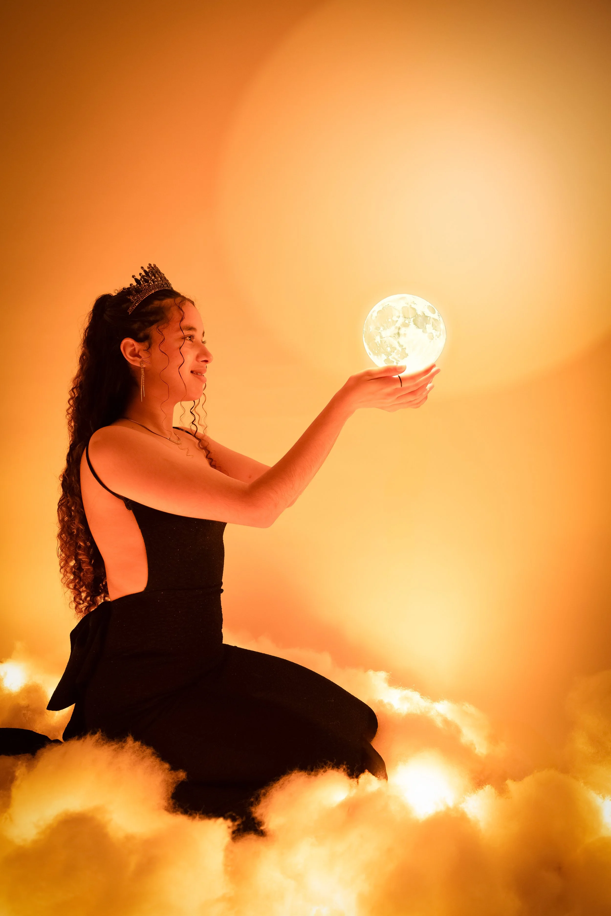 Mujer con corona y vestido negro sosteniendo la luna en la mano con fondo naranja y nubes.